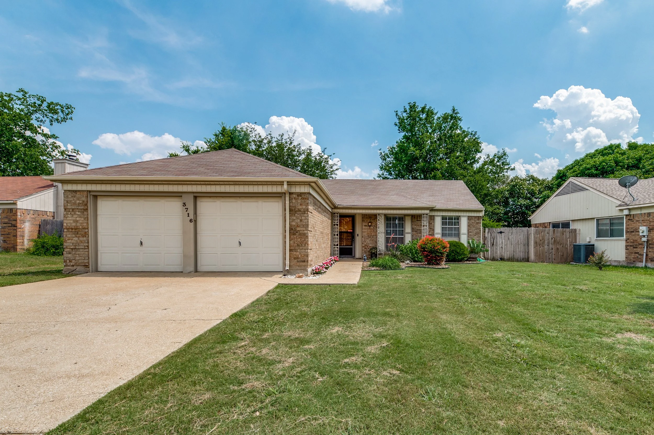 Front view of a single-story brick house with a gray-tiled roof, two garage doors, a small garden with colorful flowers, a lawn, and neighboring houses with fences under a blue sky with white clouds.