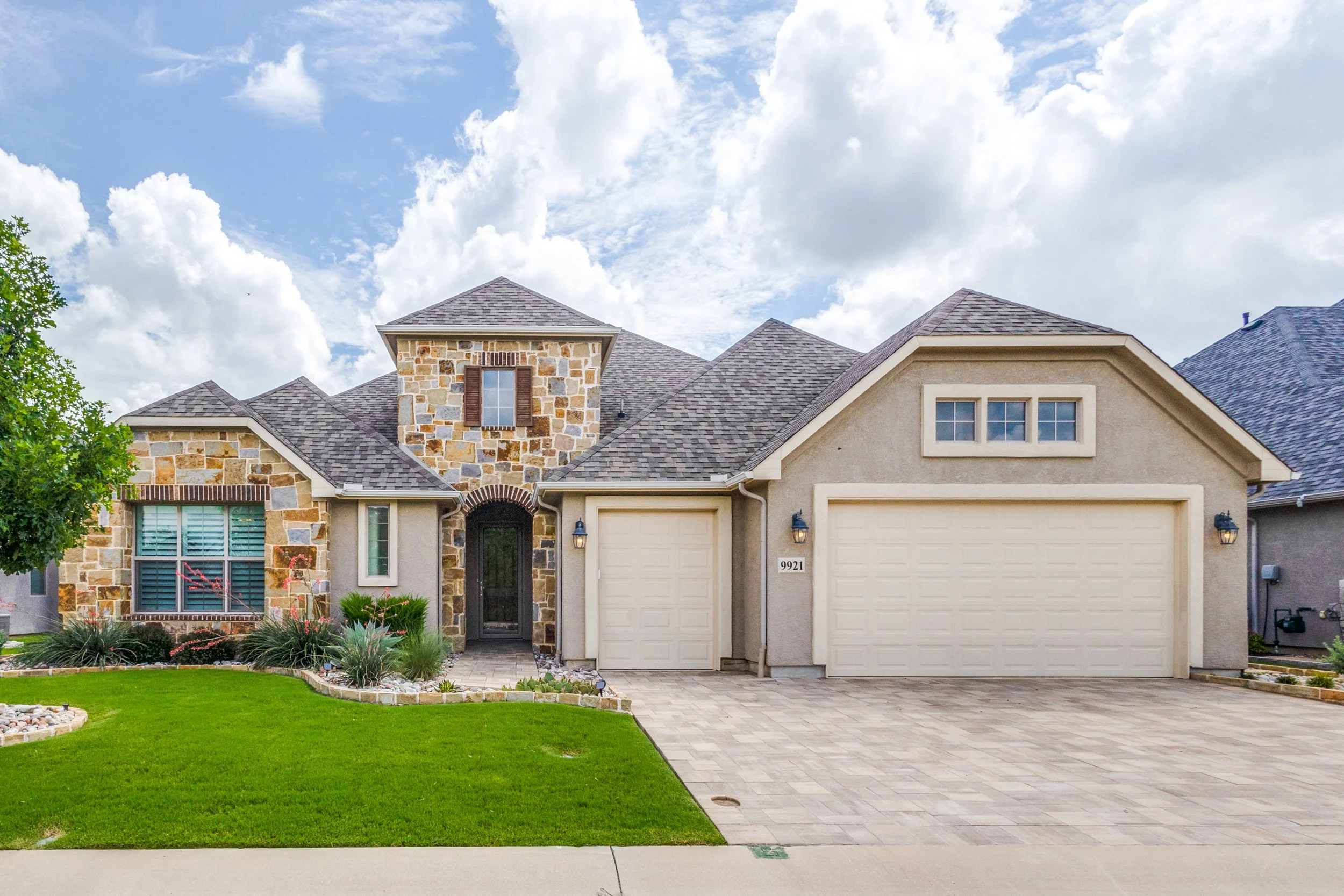 A two-story house with a stone and stucco exterior, a front lawn, a paved driveway, and a garage, under a partly cloudy sky.
