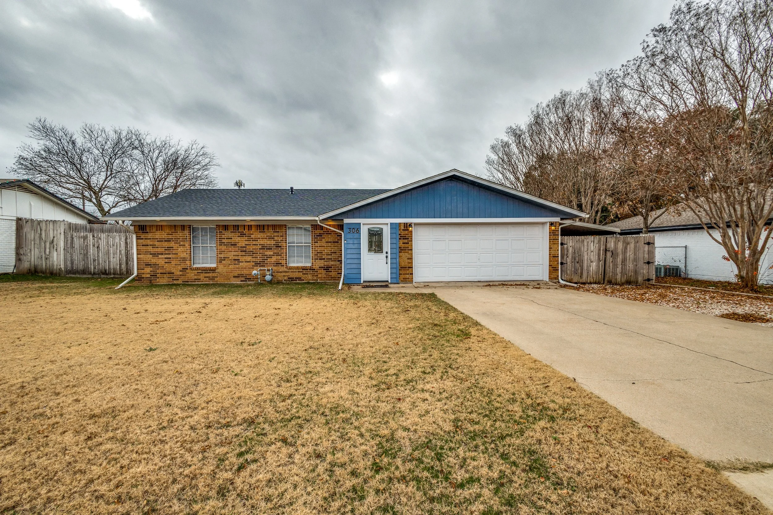 A single-story suburban house with a brick front, gray shingle roof, white garage door, blue exterior wall with house number 306, and a white front door. The yard has a dry, yellowish grass lawn and concrete driveway leading to the garage. There is a