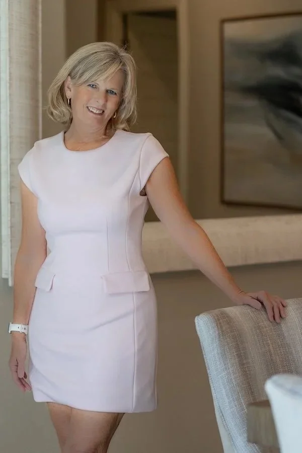 A woman with blonde hair in a white dress standing indoors, leaning on a chair.