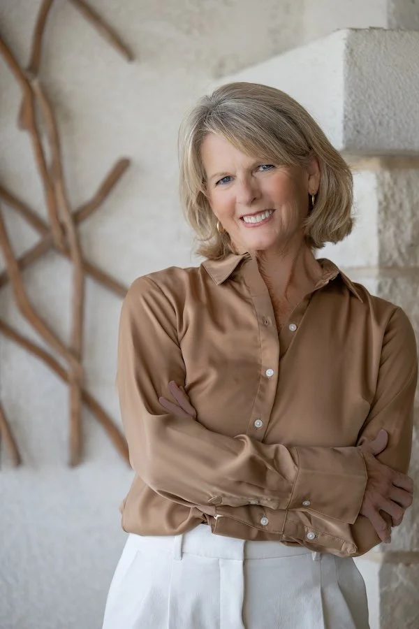A middle-aged woman with blonde hair smiling at the camera, wearing a tan satin blouse and white pants, standing with arms crossed against a decorated wall with branches.