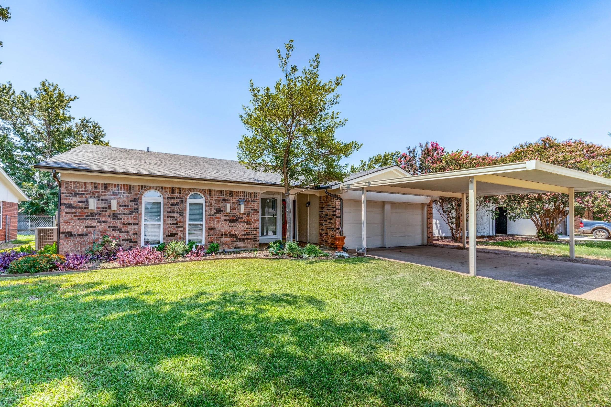 A suburban house with a brick exterior, front yard with green lawn, and a carport, surrounded by trees and plants, under a clear blue sky.