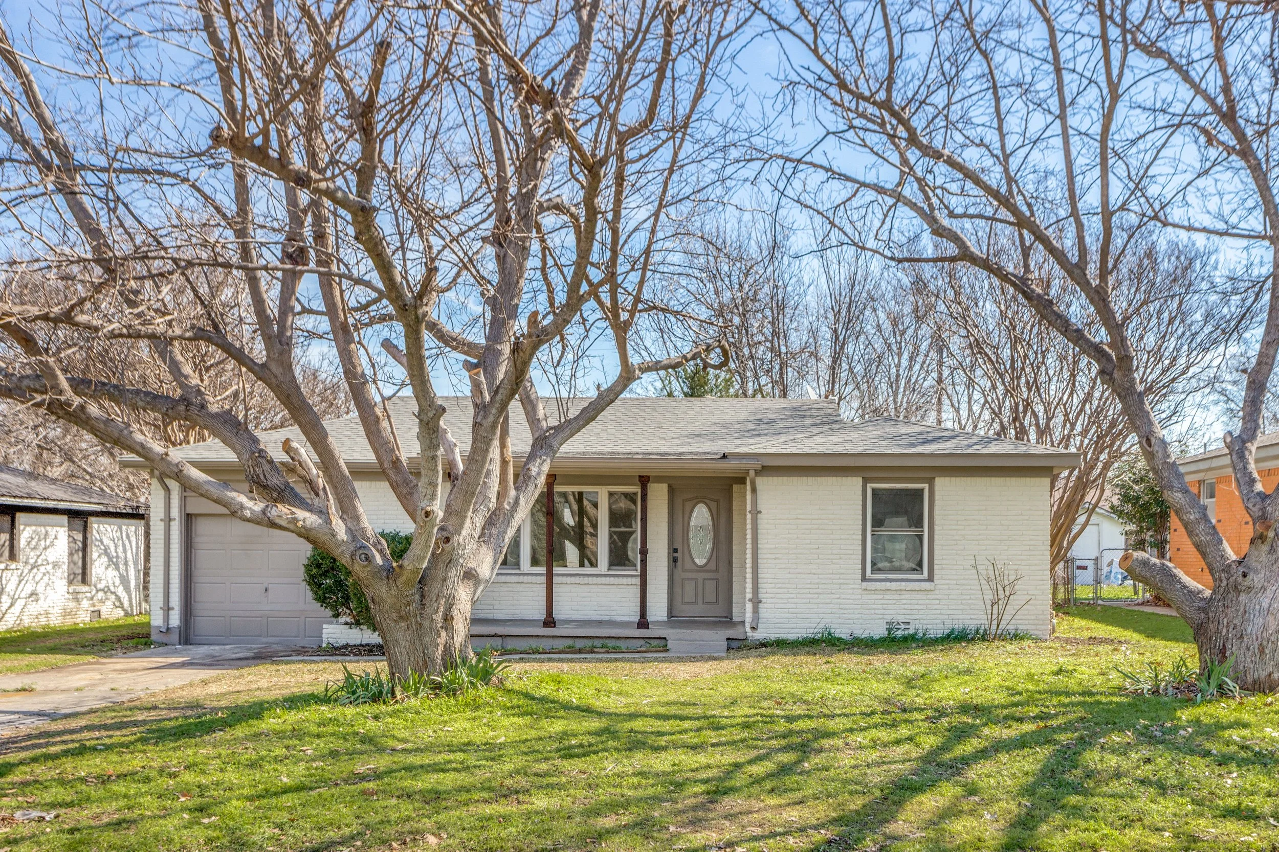 Front yard of a house with a large leafless tree, green grass, and a blue sky.