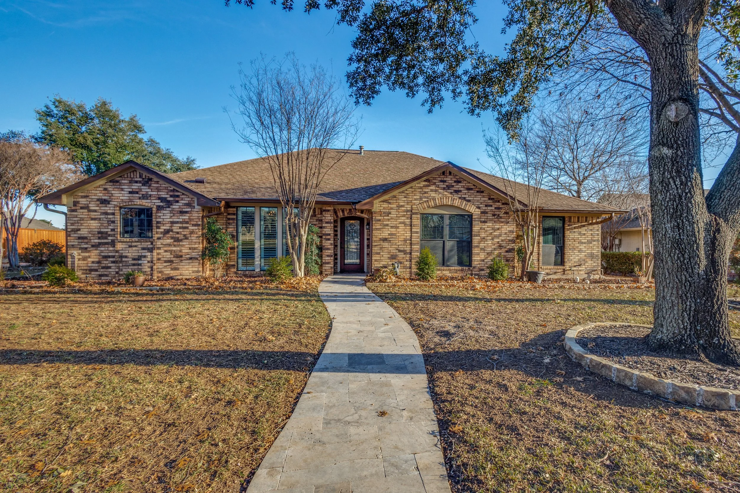 A brick house with a brown shingle roof, a front door with glass panels, and large windows. A curved concrete pathway leads to the front door. The yard has patches of brown grass and leafless trees, with some trees with green leaves. The sky is clear