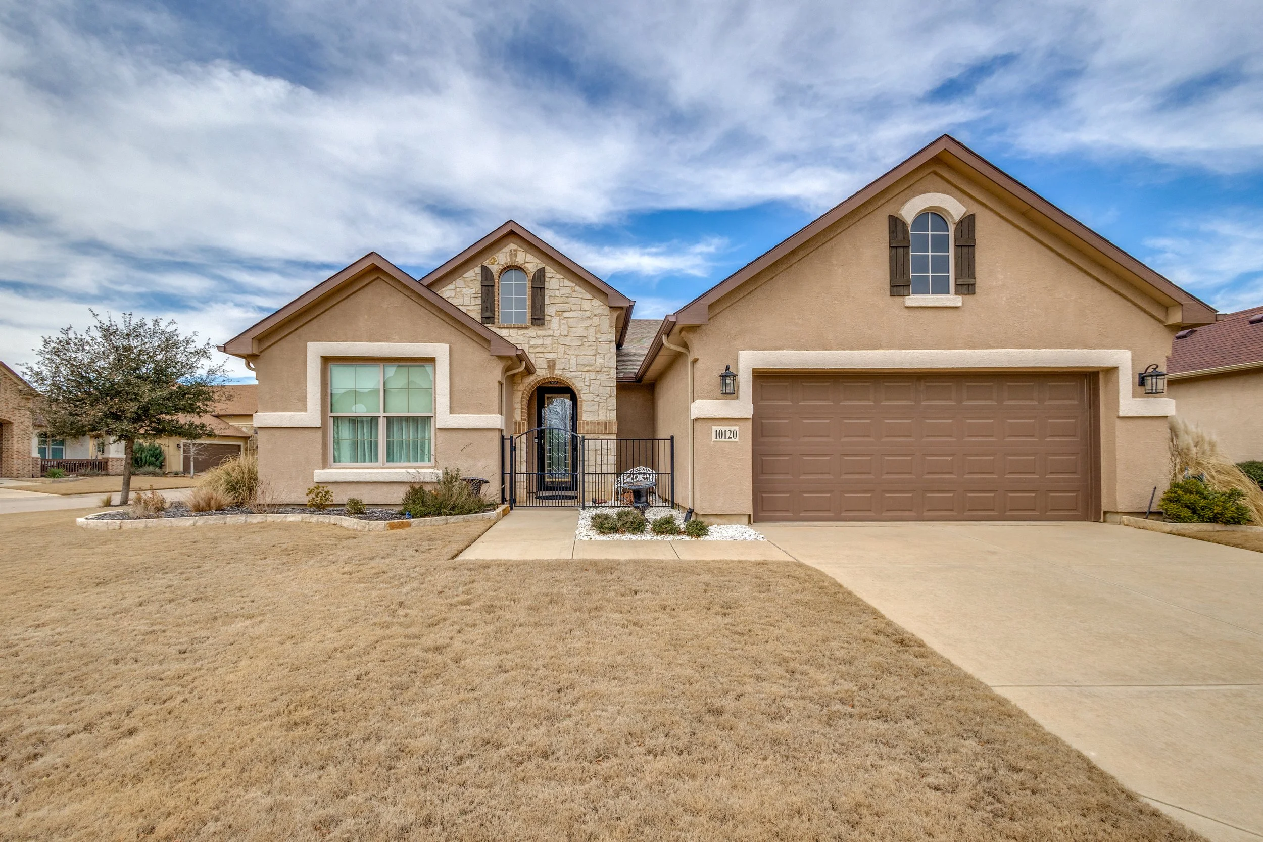 A beige house with a brown garage door, two arched windows with wooden shutters, and a front porch with a black metal gate, set against a partly cloudy sky.