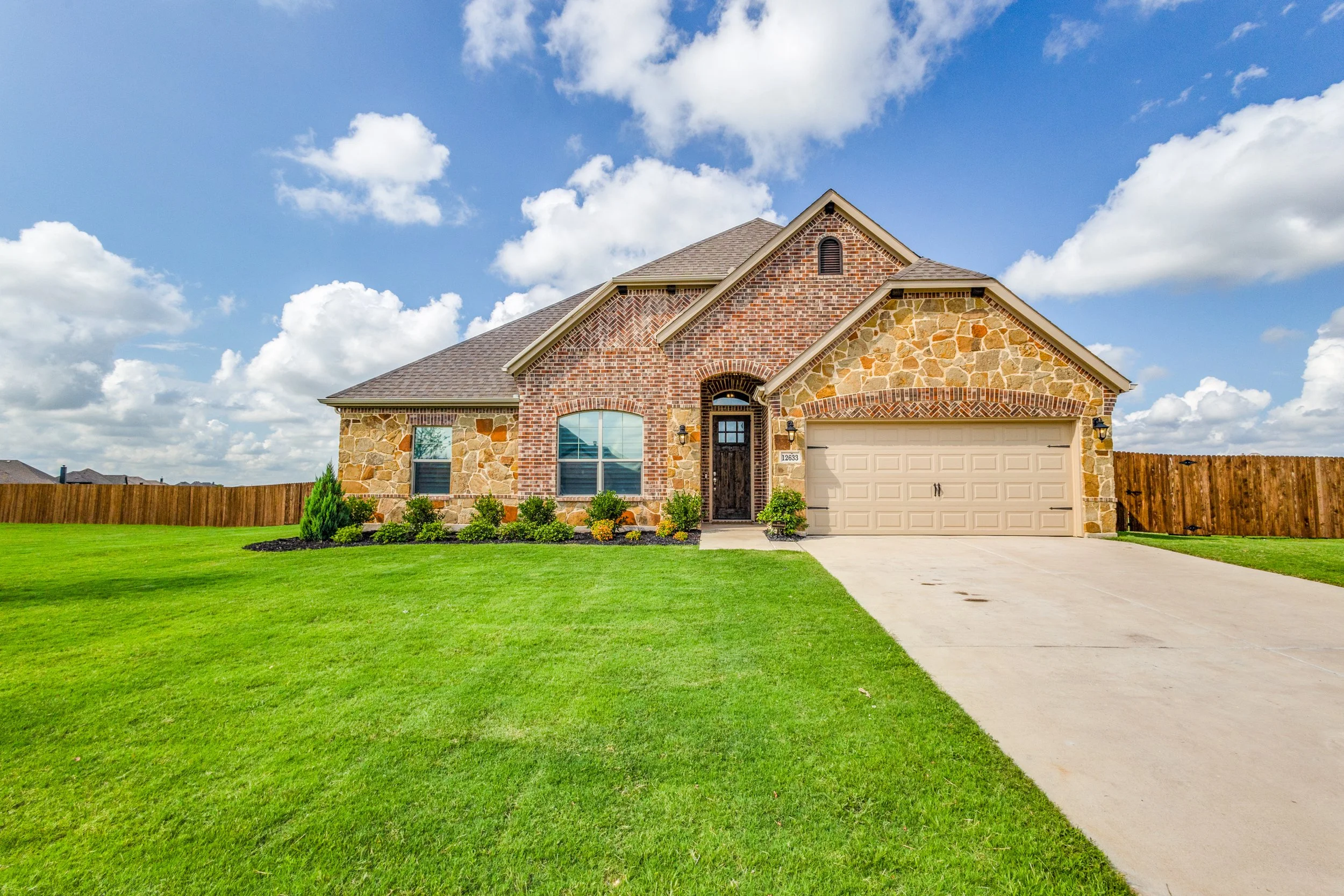 Front view of a house with a brick and stone exterior, a gray garage door, a concrete driveway, a neatly manicured green lawn, and a bright sky with scattered clouds.