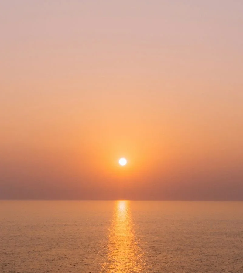 A sunset over calm ocean waters with a reflection of the sun on the water.