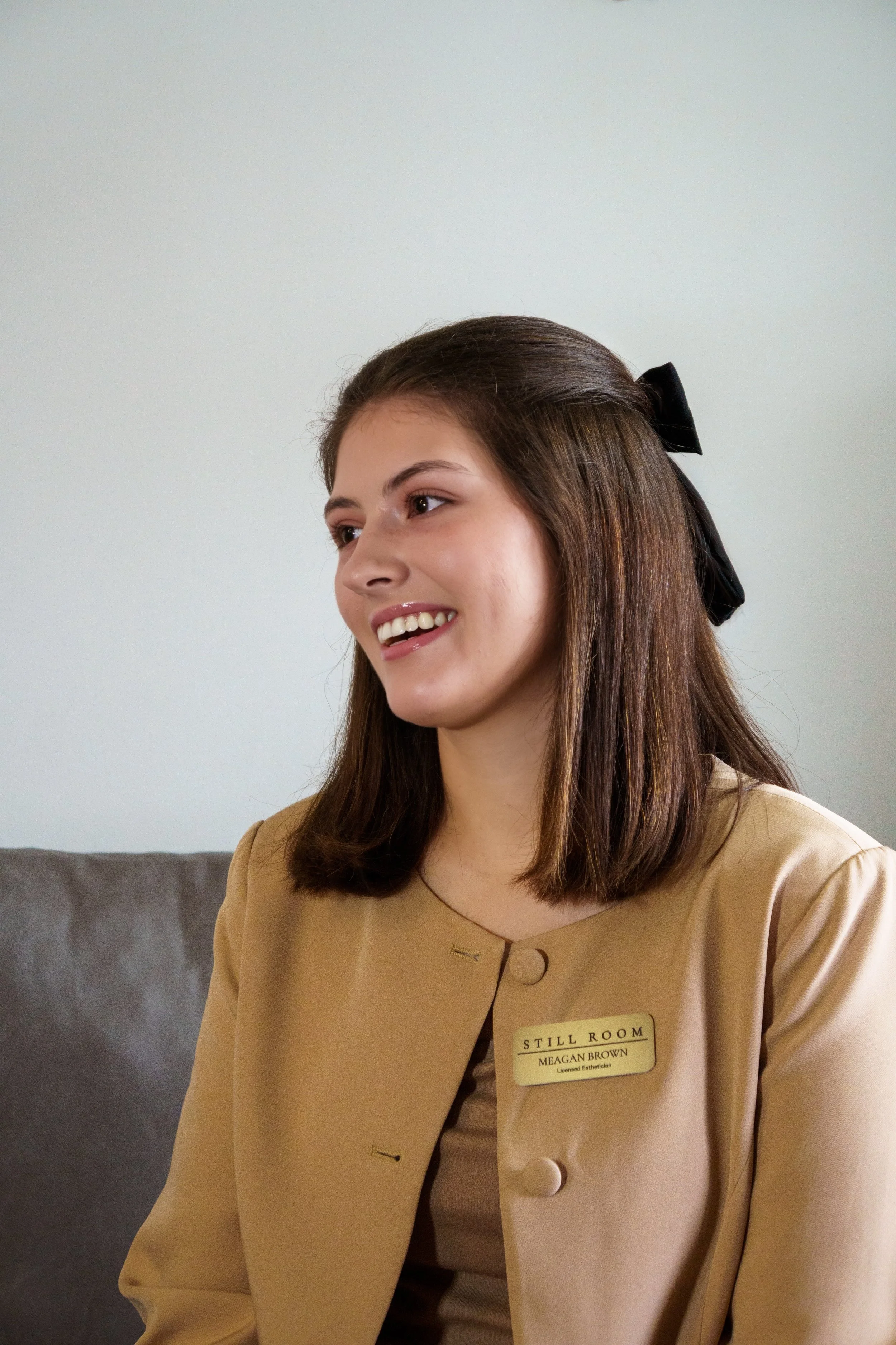A young woman with brown hair, wearing a beige uniform with a name tag that reads 'MEGAN BROWN, Licensed Esthetician,' sitting on a gray couch against a plain white wall, smiling and looking to her left.