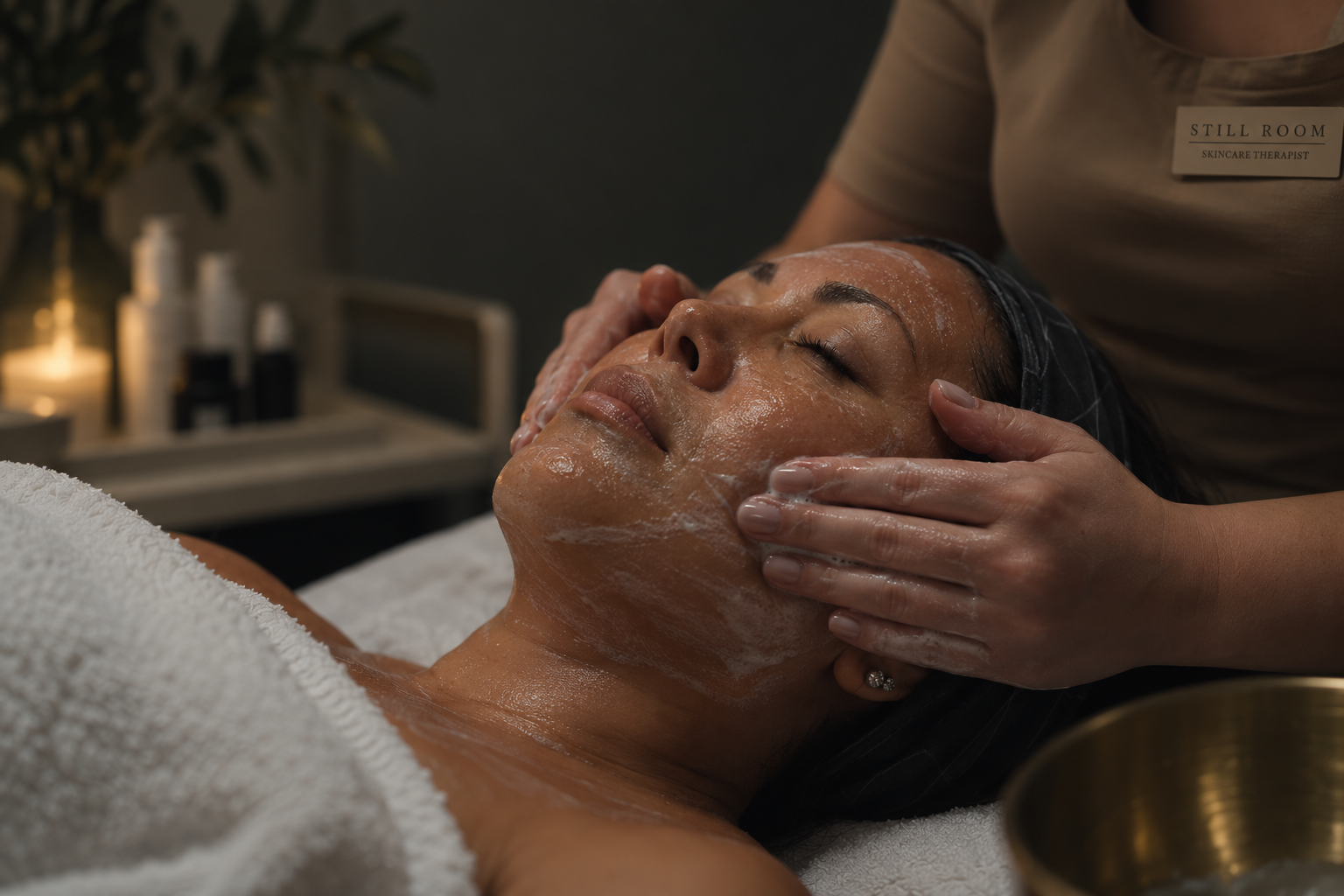A woman receiving a facial treatment, lying down with eyes closed, as a skincare therapist applies a product to her face in a spa setting.