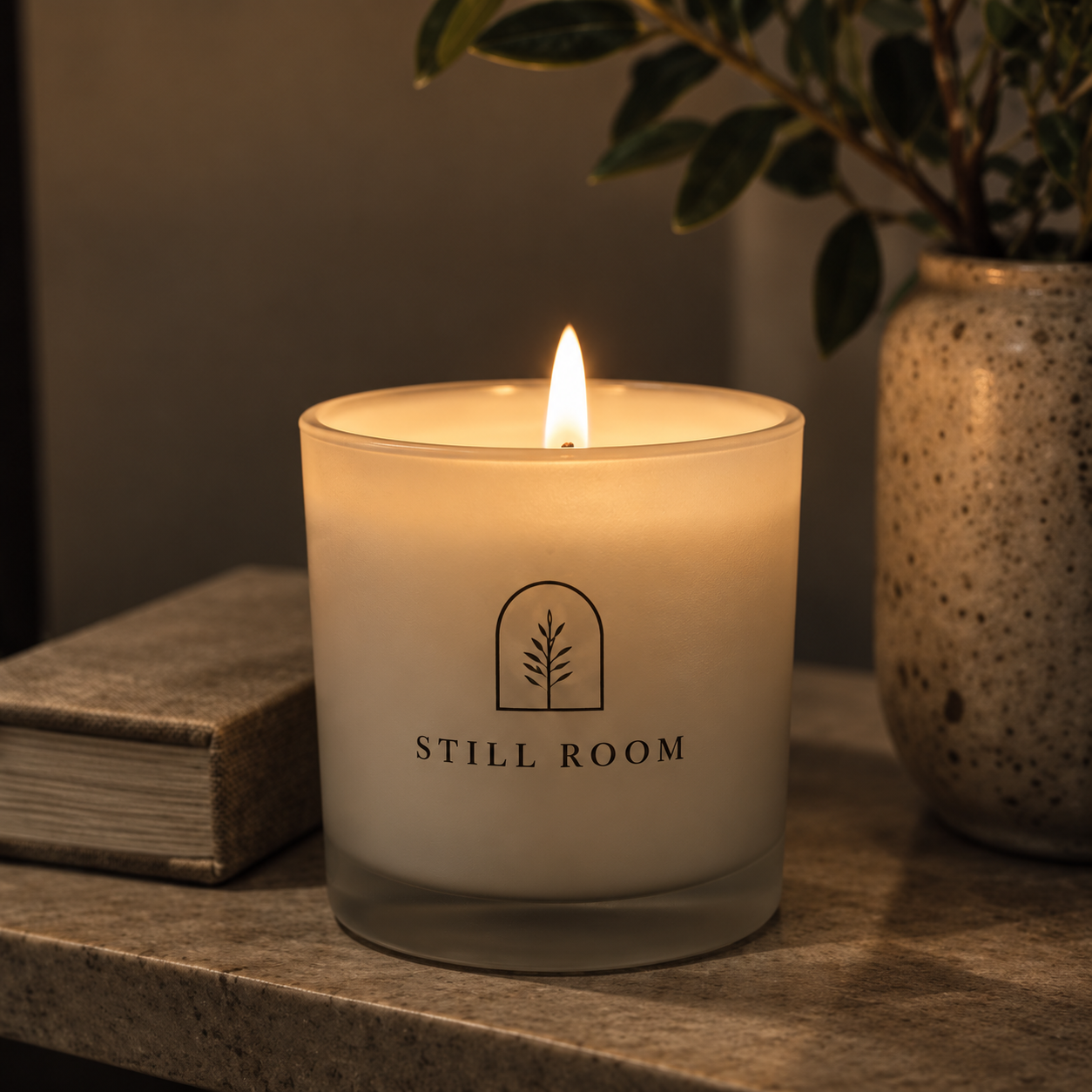 A lit white candle with the label 'Still Room' placed on a tabletop next to a closed book and a speckled ceramic vase with green leafy plant.