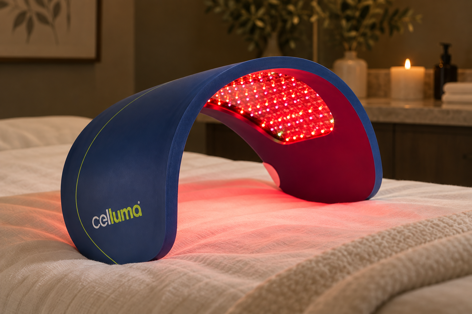 A blue and red LED light therapy device with the brand name 'Celluma' on it, resting on a bed in a bedroom with candles and houseplants in the background.