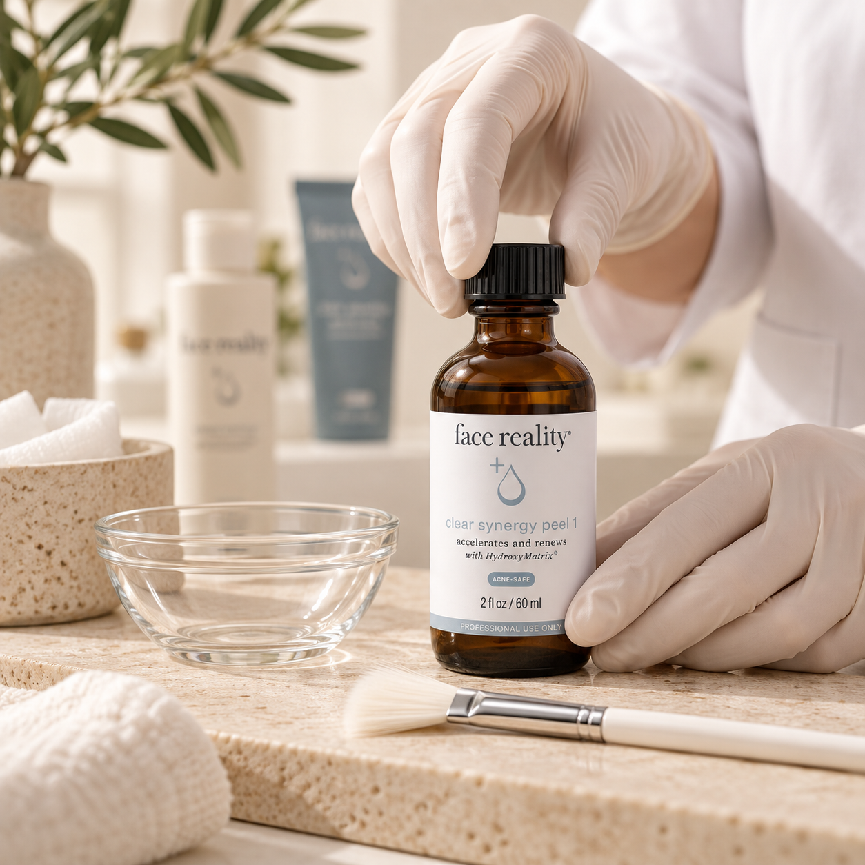 Person wearing gloves handling a brown bottle labeled 'face reality' skincare product on a light-colored counter, with skincare items, a glass bowl, a brush, and a towel nearby.