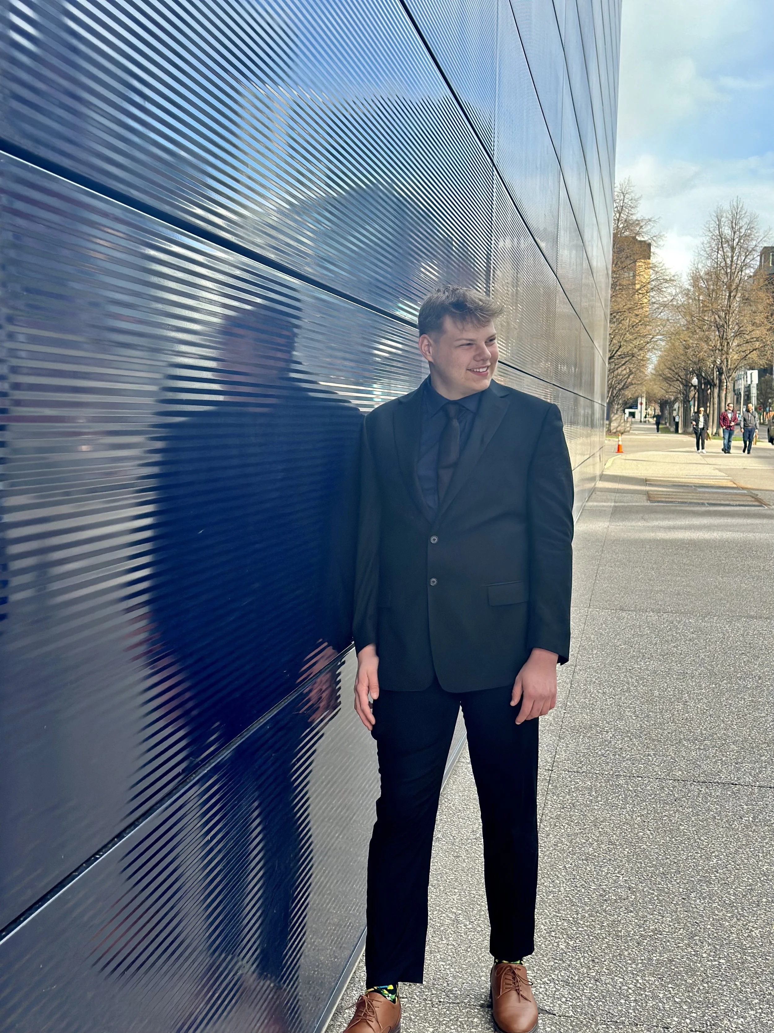 A young man in a black suit and tie standing outdoors next to a modern, metallic building with horizontal ridges. The young man is smiling and looking to his right, wearing brown shoes and colorful socks. There are trees and a few pedestrians in the background.