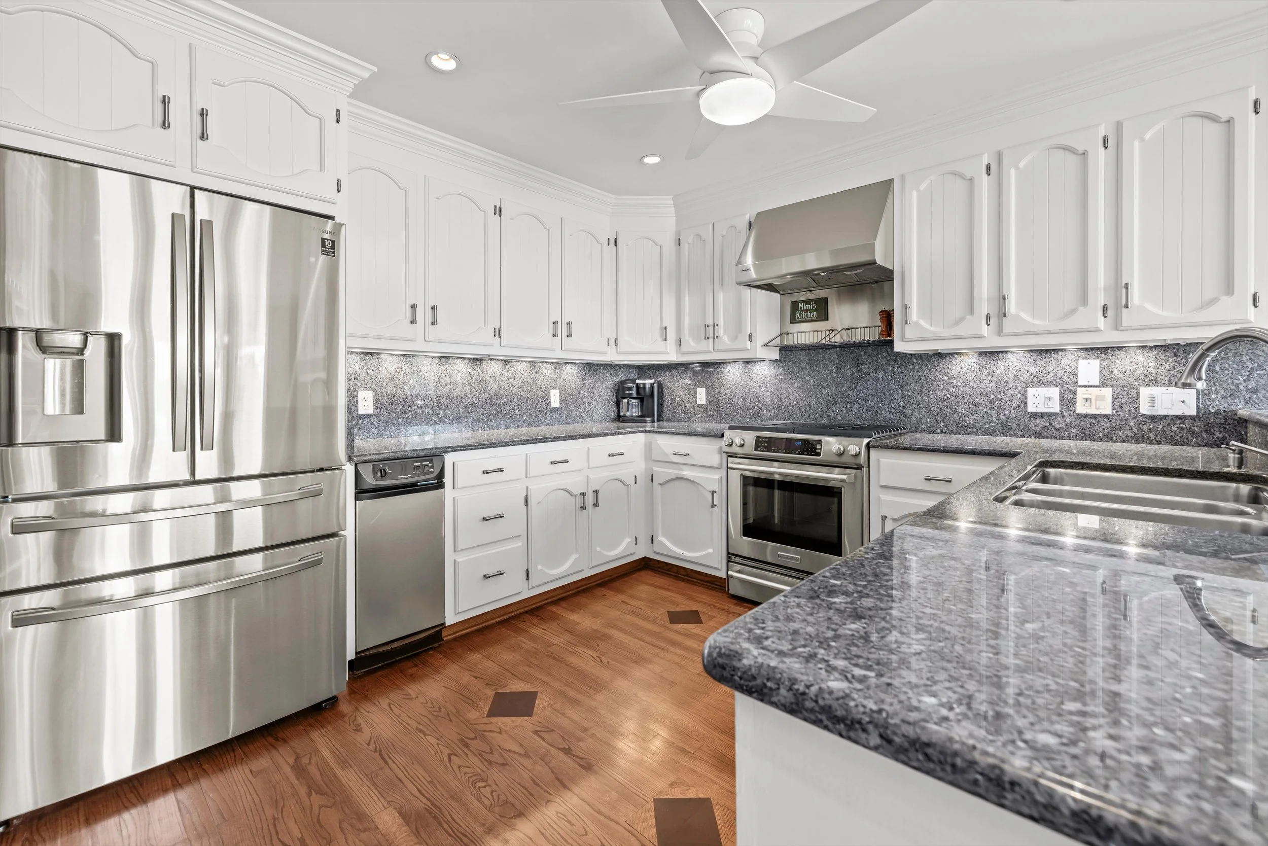 A modern kitchen with white cabinets, stainless steel appliances, granite countertops, hardwood floors, and a ceiling fan.