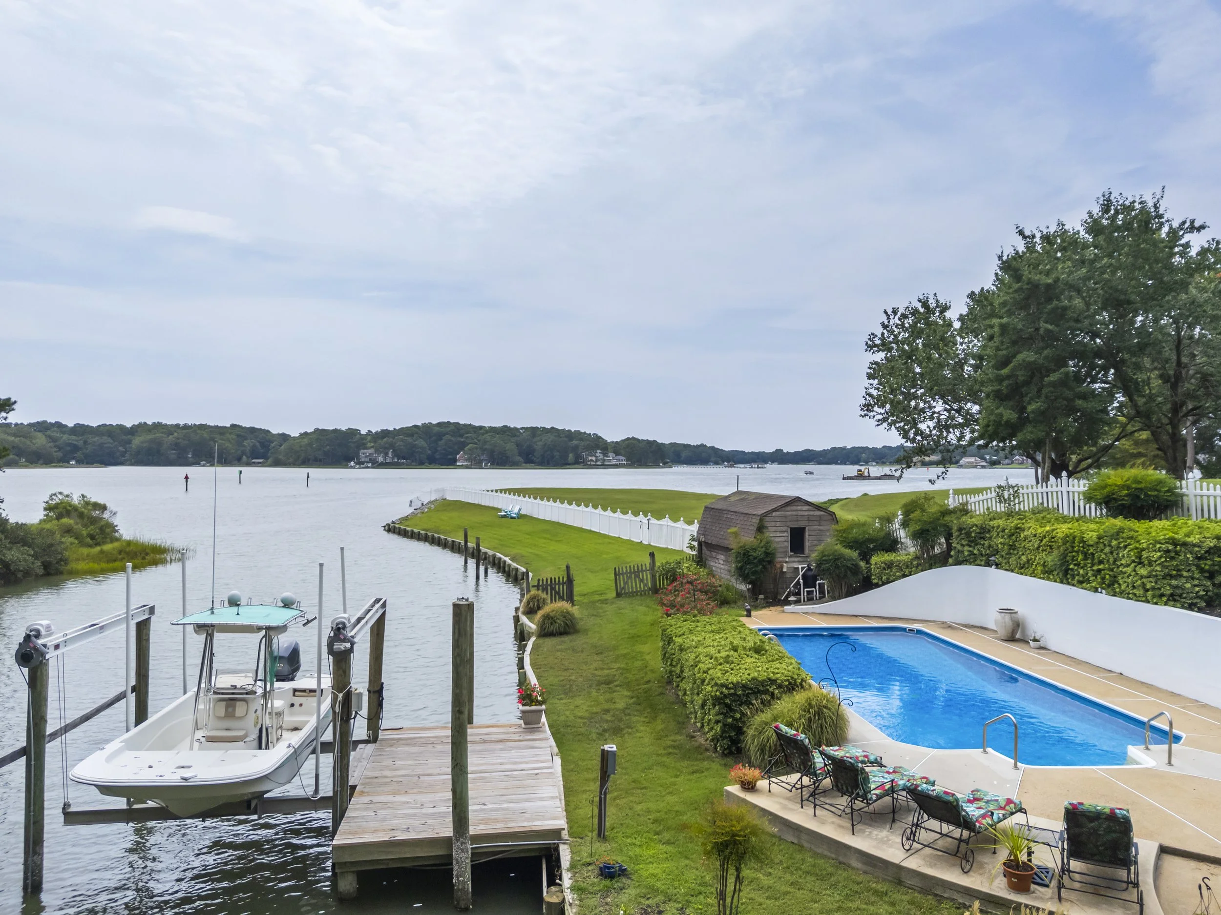 View of a backyard with a swimming pool, outdoor furniture, a small wooden shed, and a boat docked at the pier, overlooking a large body of water with trees in the background.