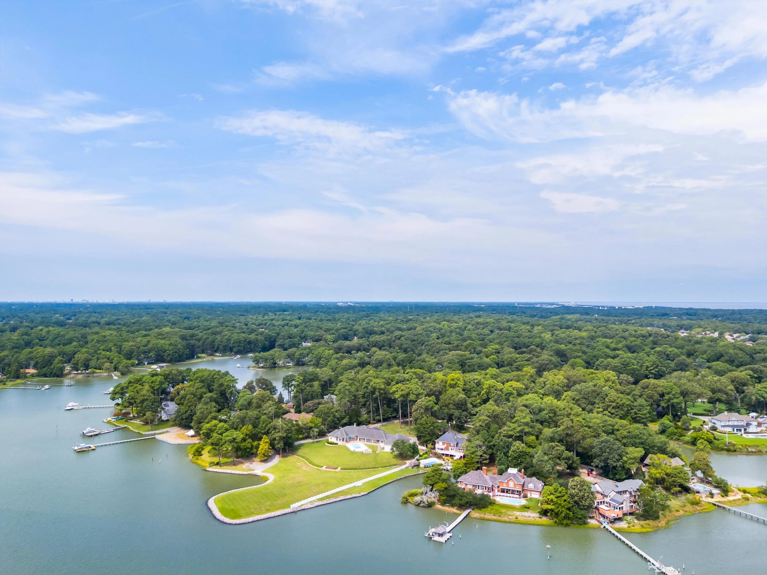 Aerial view of a lakefront neighborhood with houses, docks, and lush green trees, under a bright blue sky with scattered clouds.
