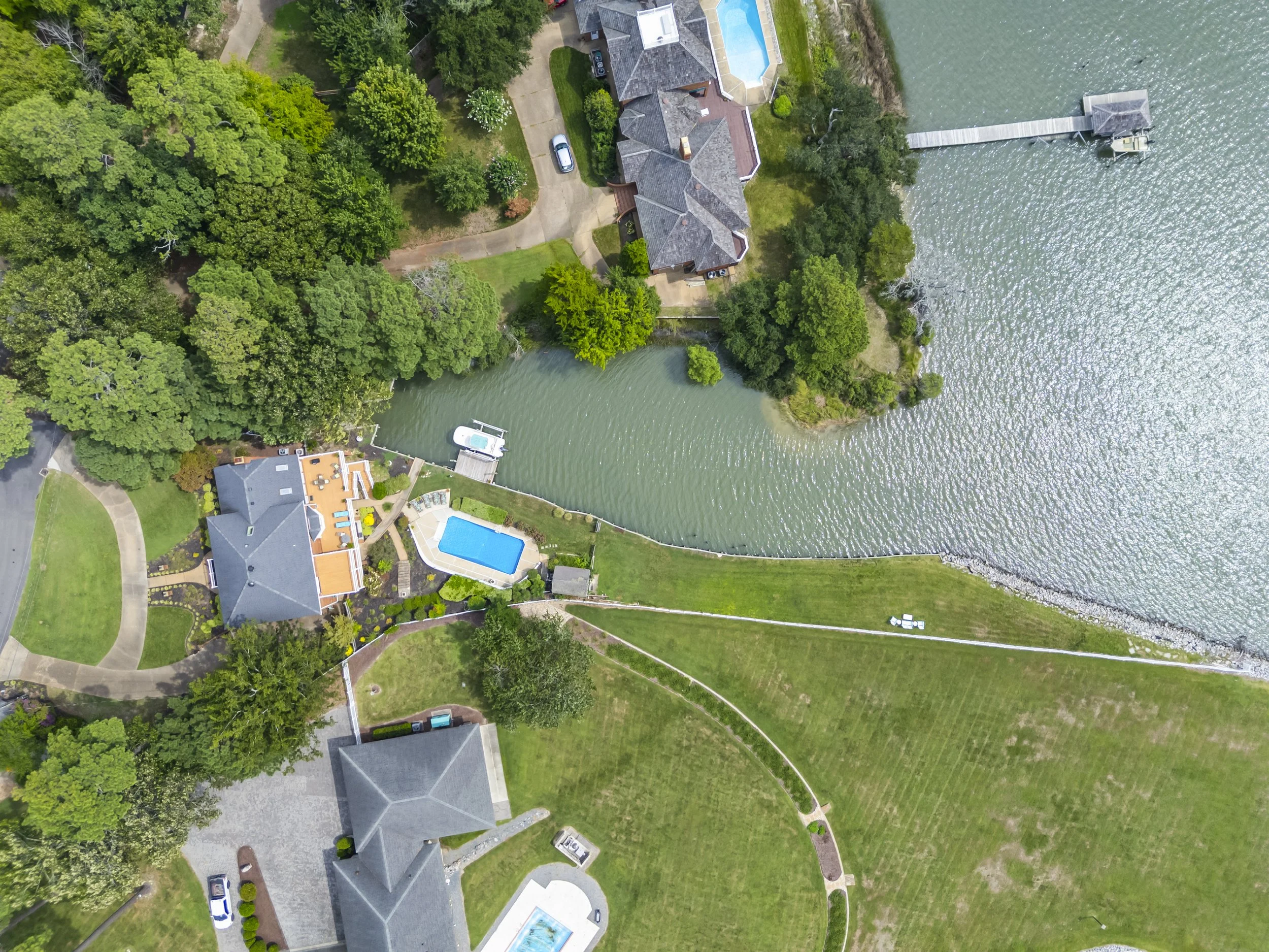 Aerial view of houses with swimming pools, lush green trees, and a waterfront with a pier.