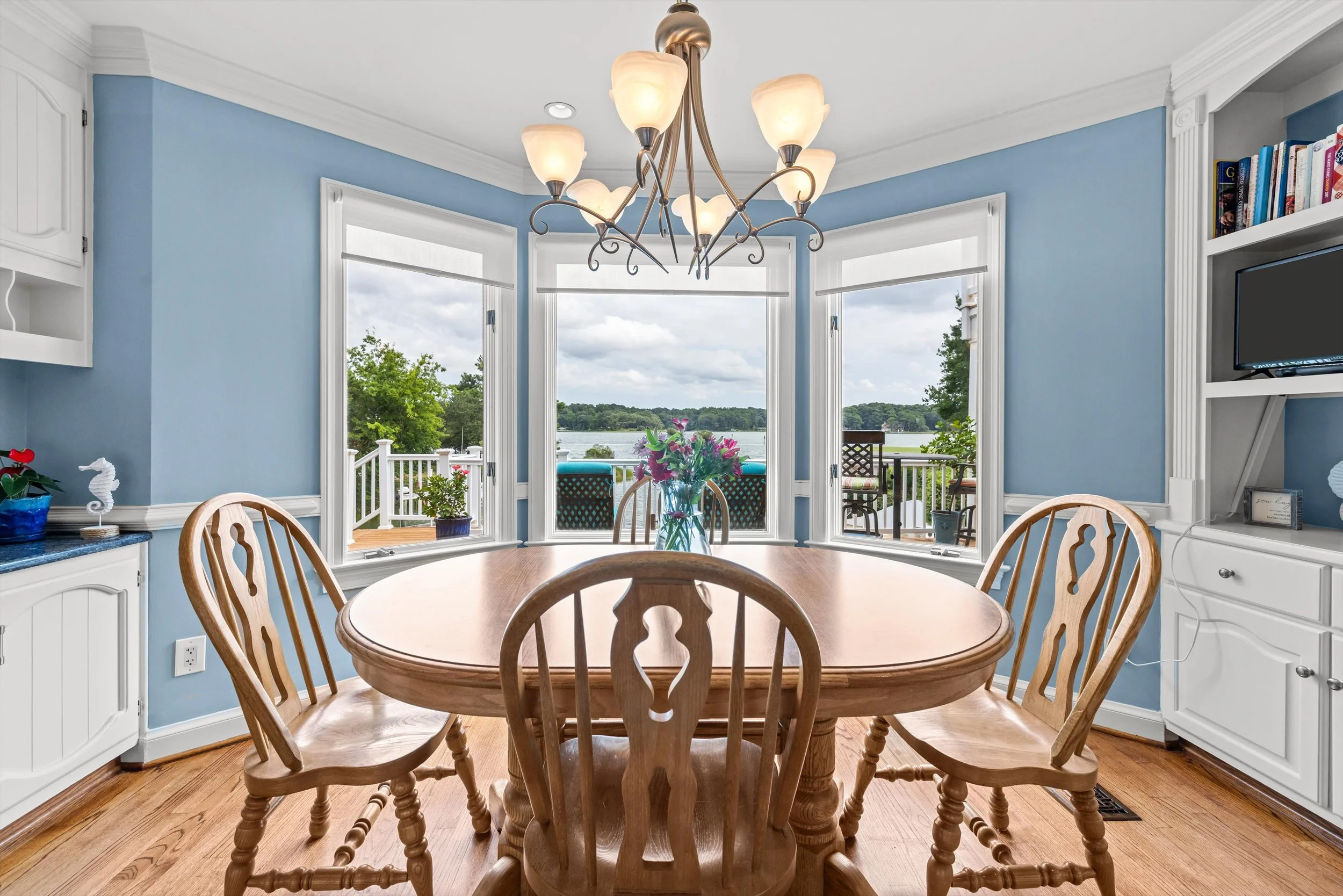 A dining room with a wooden table and matching chairs, blue walls, large windows overlooking water and trees, a chandelier hanging from ceiling, and built-in shelves with books and a TV.