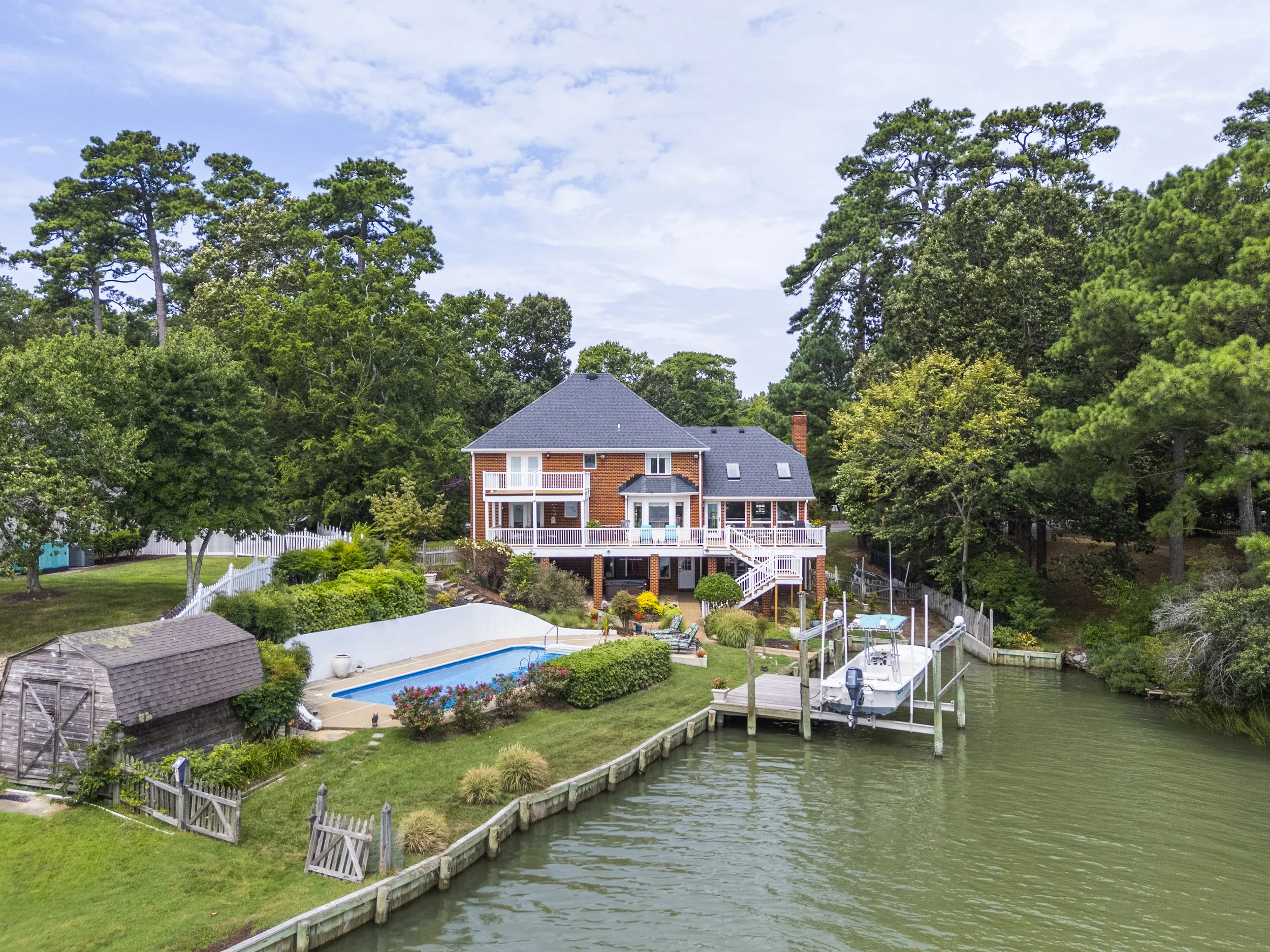 A two-story brick house with a gray roof, white porch railings, and multiple windows, situated in a lush green yard with trees and a swimming pool. The house is next to a canal with a dock and a boat, surrounded by a white picket fence.