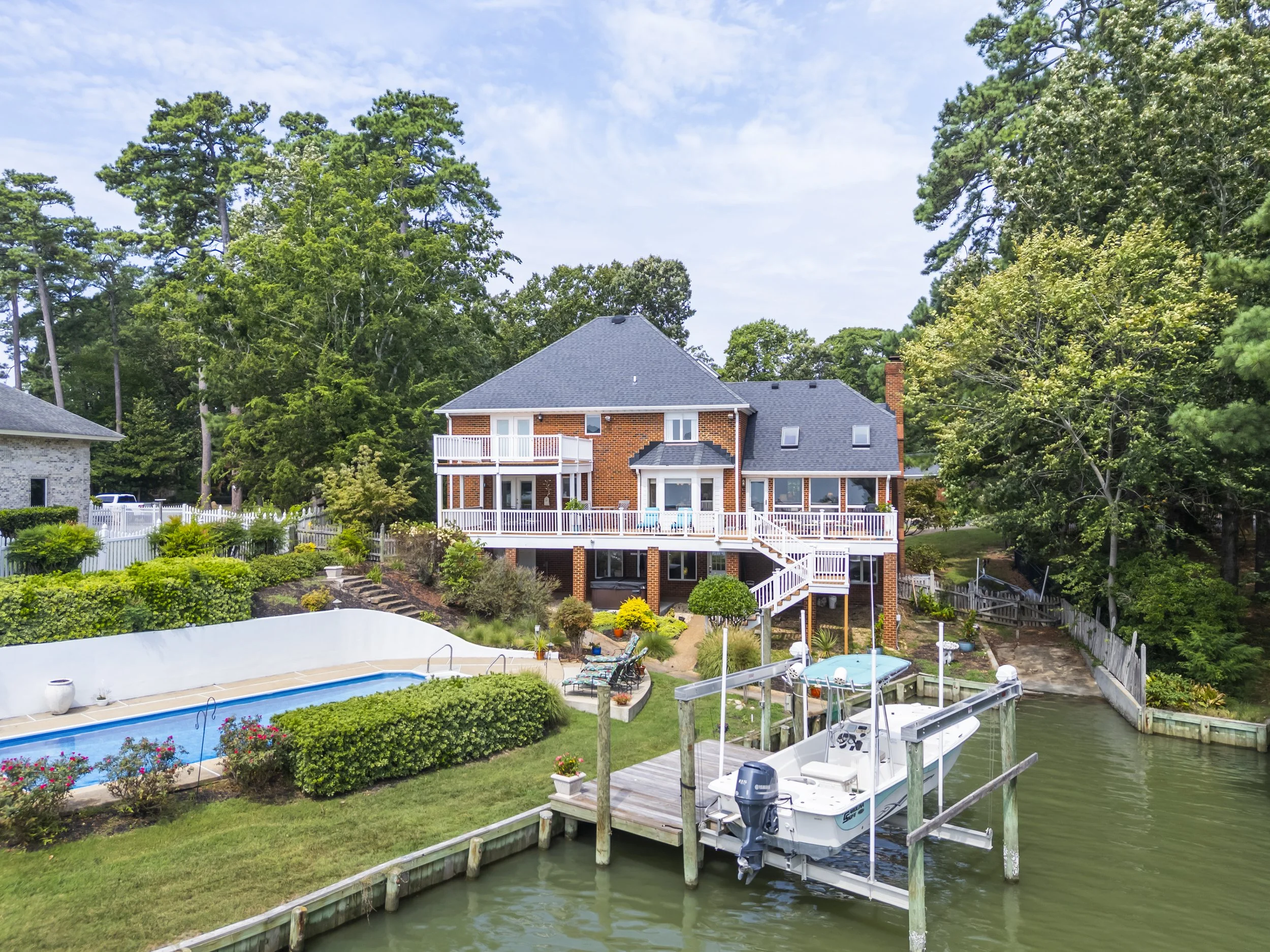 A waterfront backyard with a swimming pool, a dock with a boat, and a brick house in the background surrounded by tall trees and greenery.