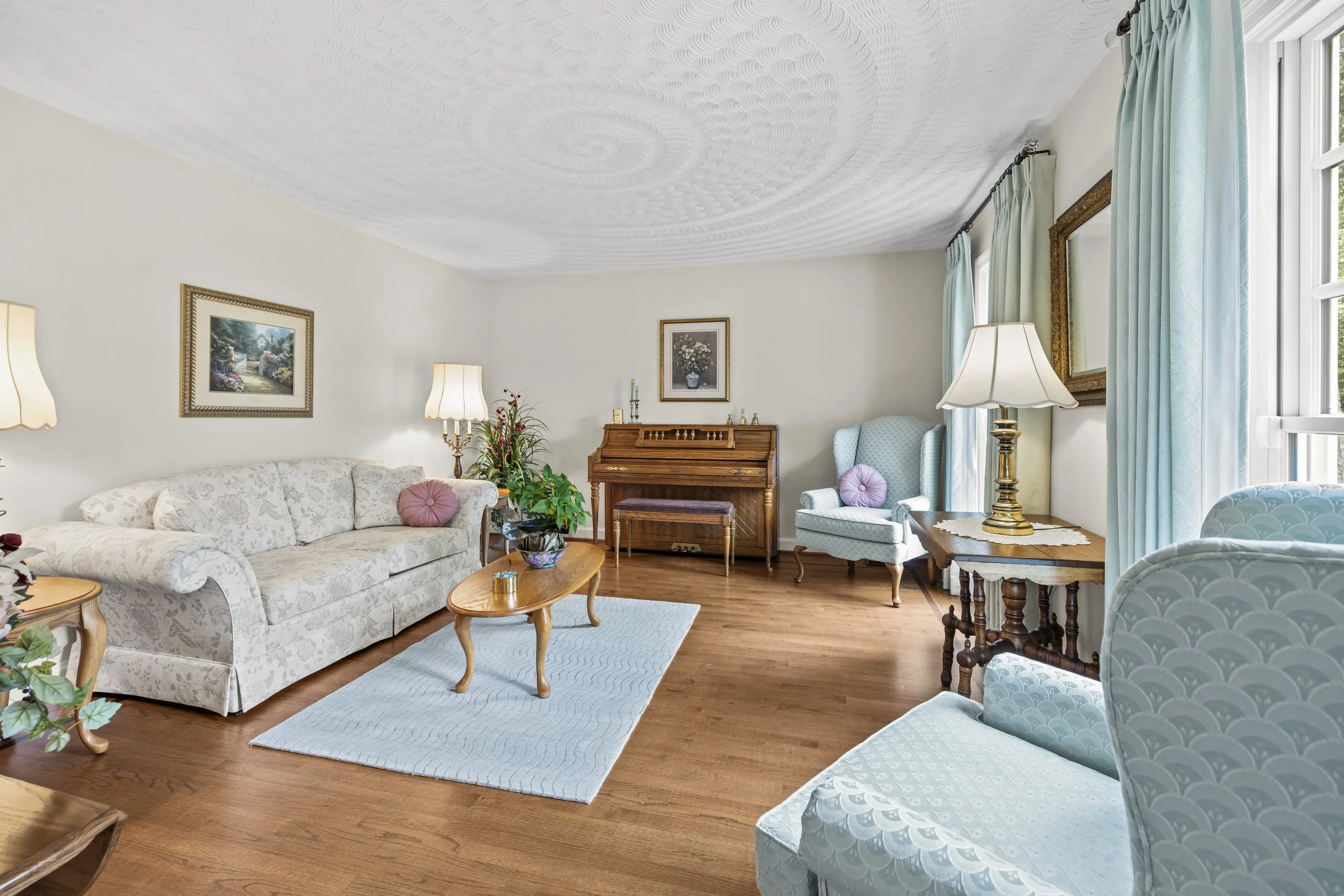 A living room with a white floral-patterned sofa, a wooden coffee table, a small round side table, a light blue armchair with a purple pillow, a wooden piano, framed paintings, a plant, a lamp, and large windows with blue curtains.