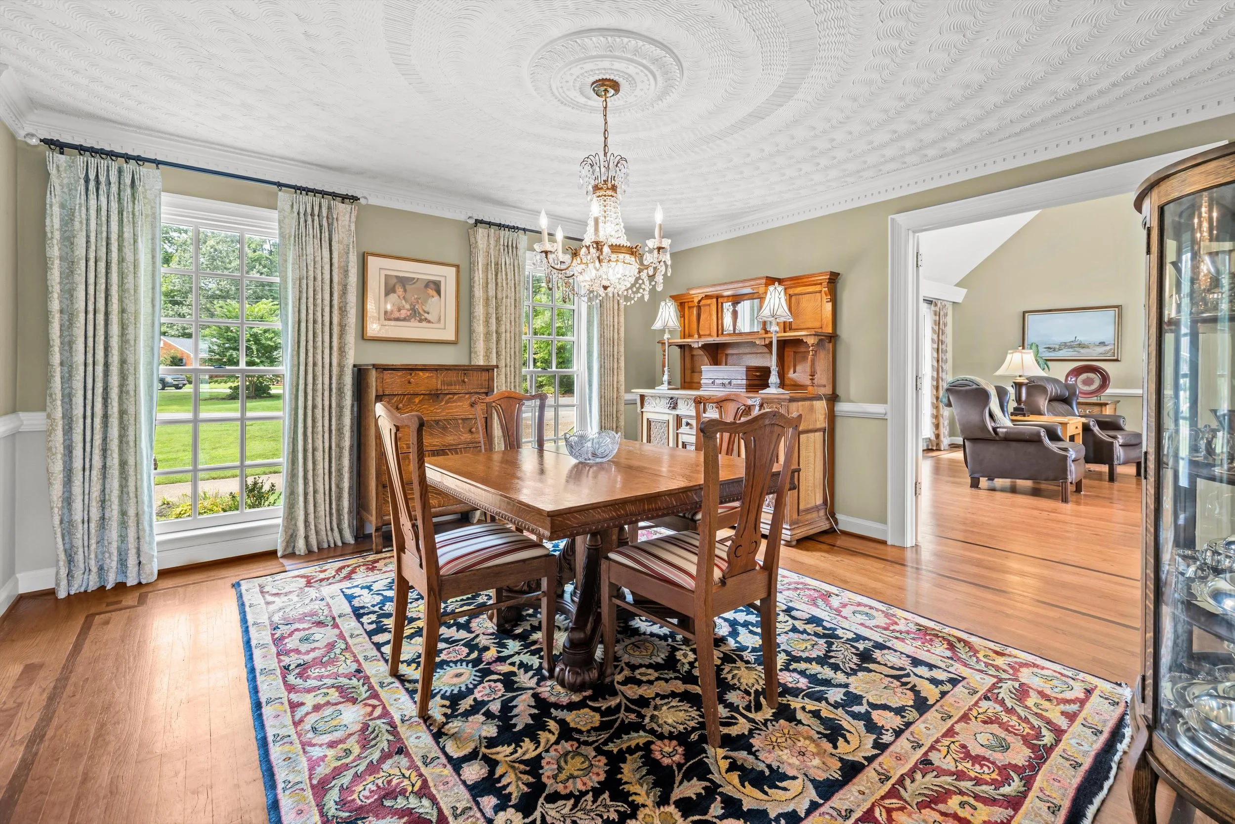 A dining room with a wooden dining table, six chairs with striped cushions, a chandelier, large windows with patterned curtains, a colorful area rug, and a wooden cabinet.