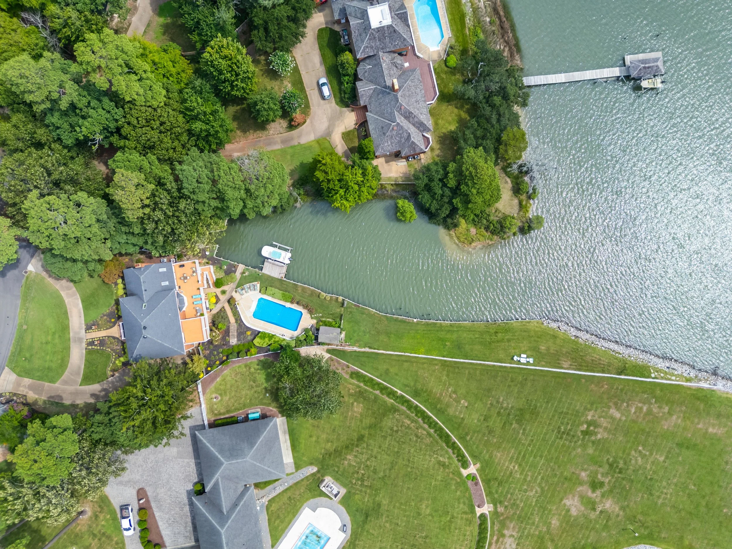 Aerial view of a lakeside residential property with a house, swimming pool, trees, lawns, and a dock extending into the water.