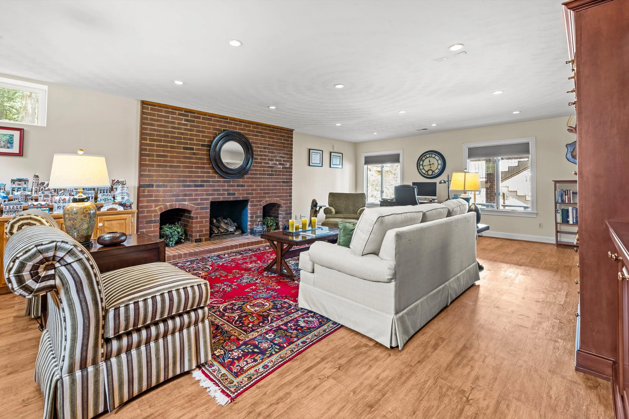 Living room with brick fireplace, patterned rug, beige sofa, striped armchair, wooden furniture, windows with blinds, and decor like framed art, a clock, and lamps.