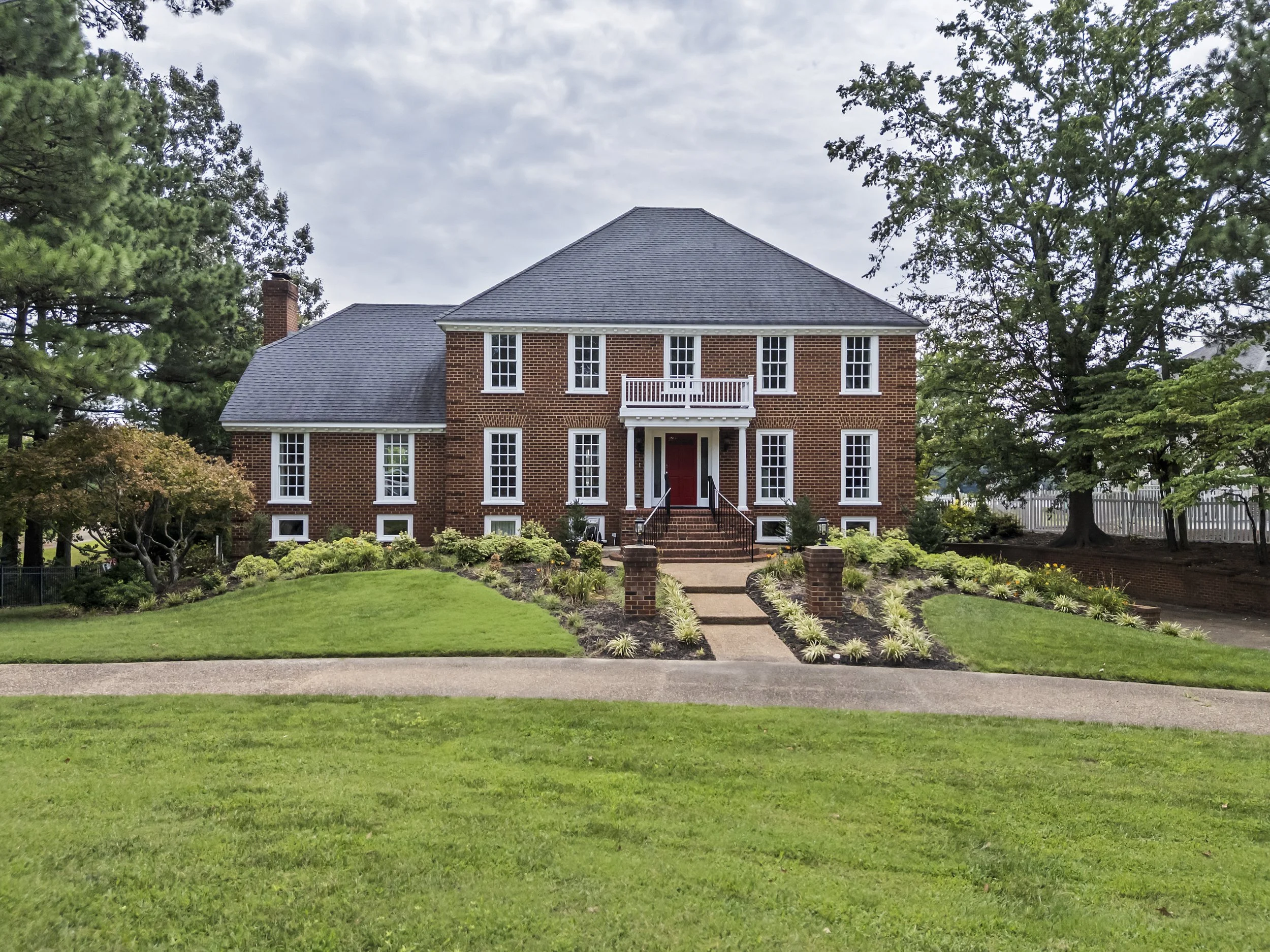 A large red brick house with white window frames, a red front door, and a small balcony above the entrance. The house is surrounded by a well-maintained garden with green grass, shrubs, and trees. The sky is overcast.