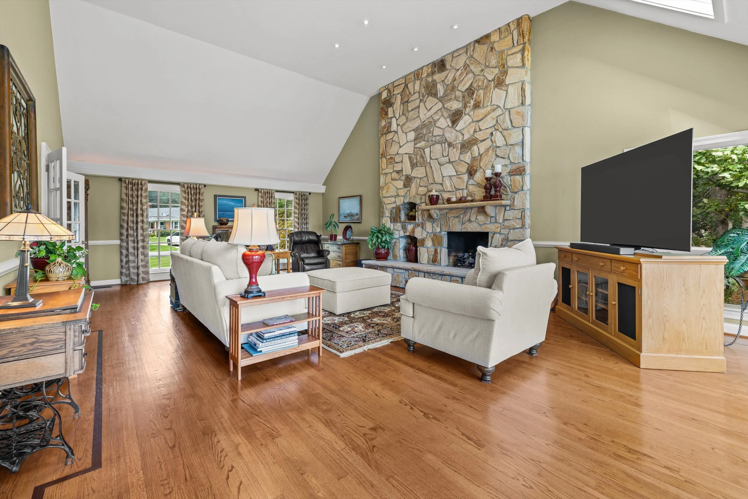 Living room with a stone fireplace, cream-colored sofas, a wooden cabinet with a television, a patterned area rug, and large windows with patterned curtains, hardwood floors, and a ceiling with recessed lighting.