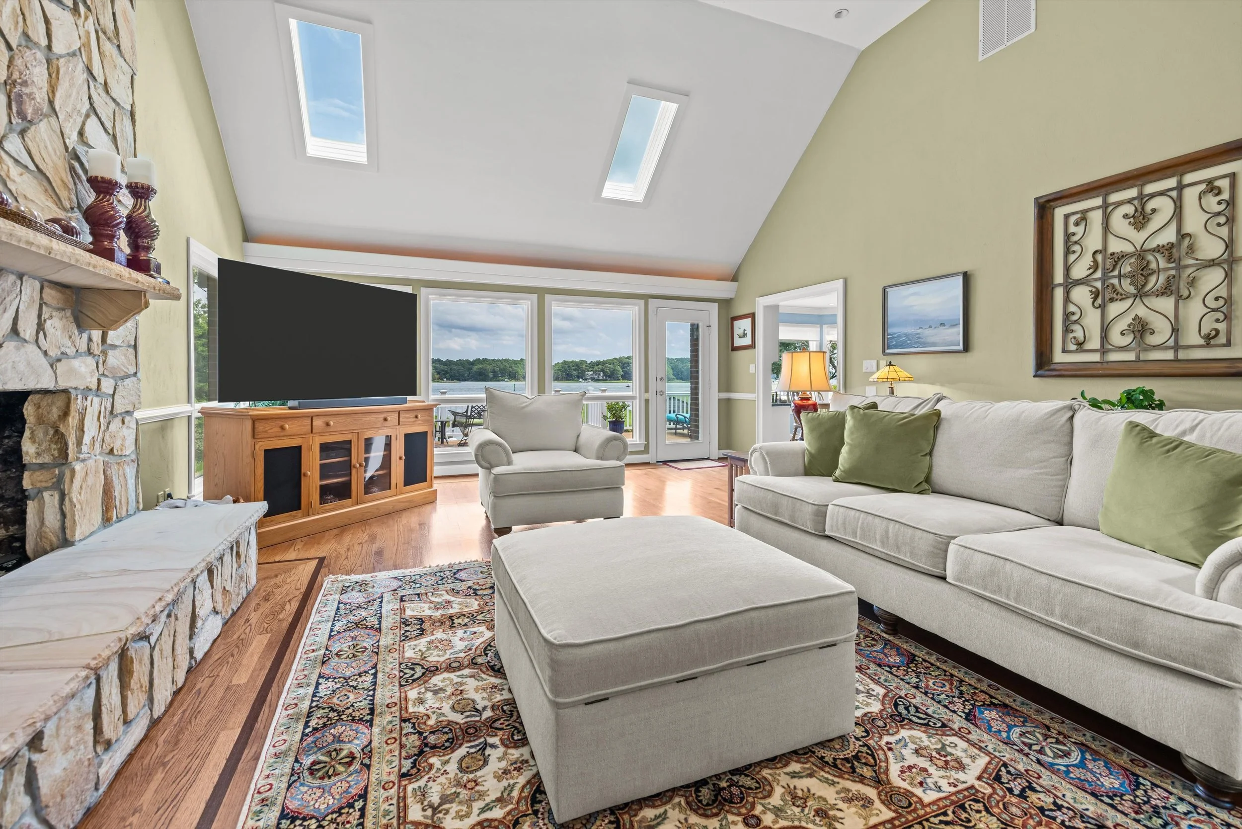 Living room with beige sectional sofa, white armchair, and colorful patterned rug. Stone fireplace with candles, large windows and glass door overlooking a lake. Walls painted light yellow and white ceiling with skylights. Art and decor on walls, pot