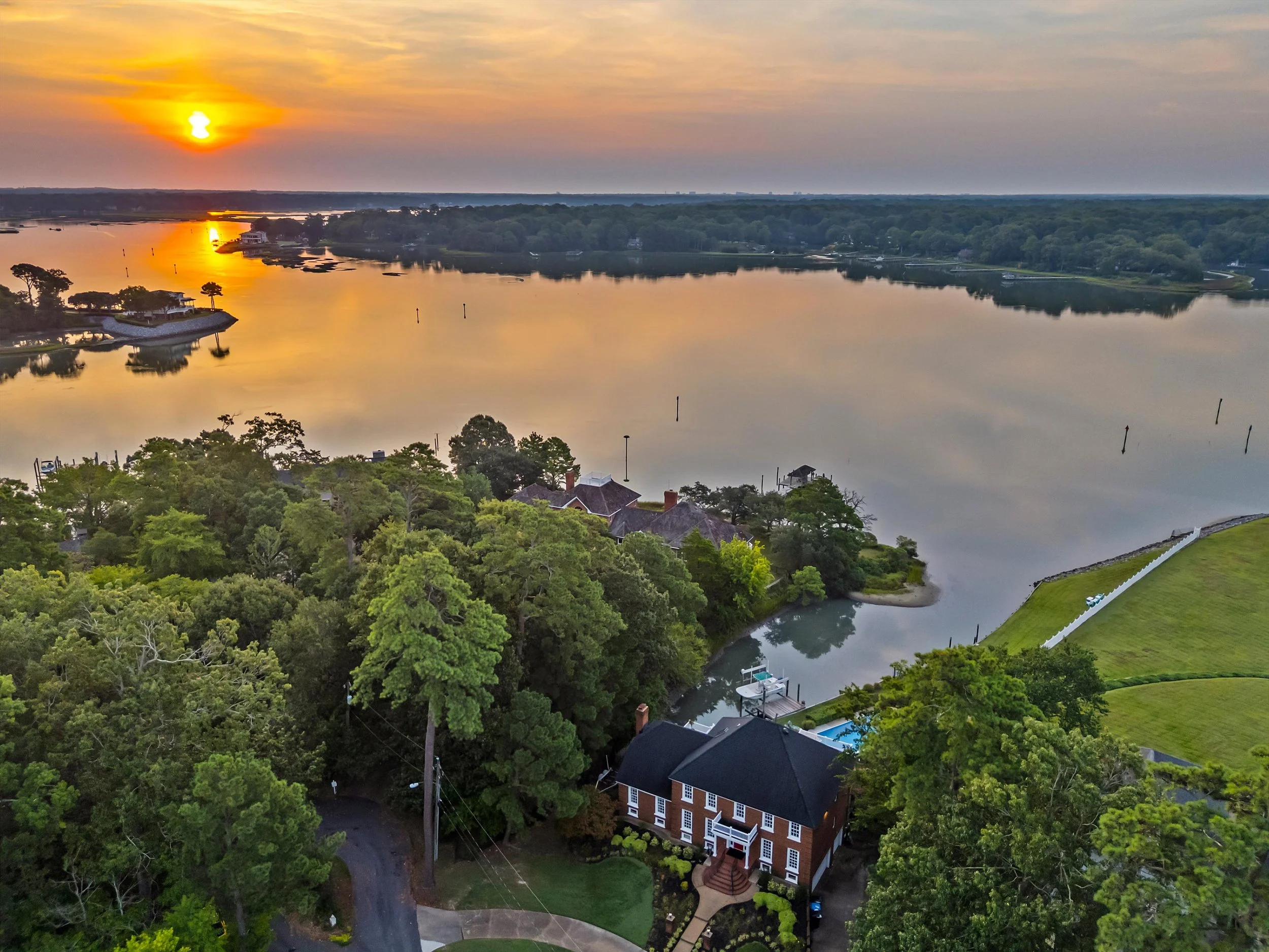 An aerial view of a river during sunset, with reflections of the setting sun on the water. Houses are surrounded by lush green trees along the riverbank, and a large house with a dark roof and a porch is visible in the foreground.