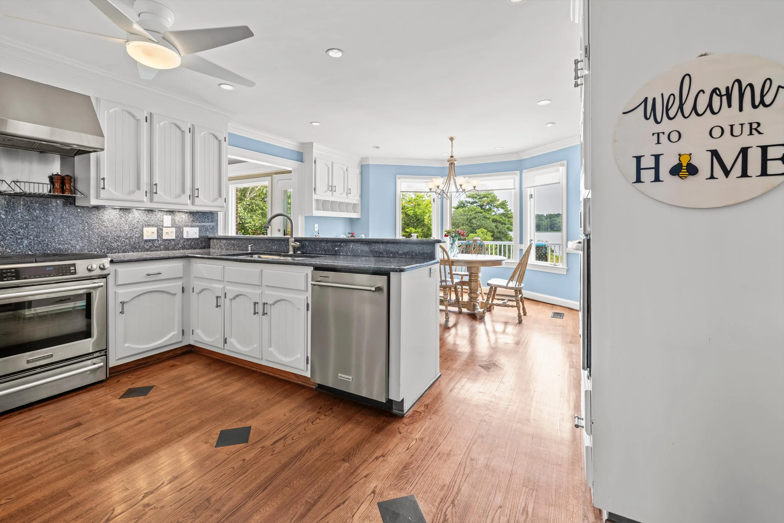 Bright kitchen with white cabinets, black granite countertops, and a wooden dining table with chairs by large windows showing green trees.