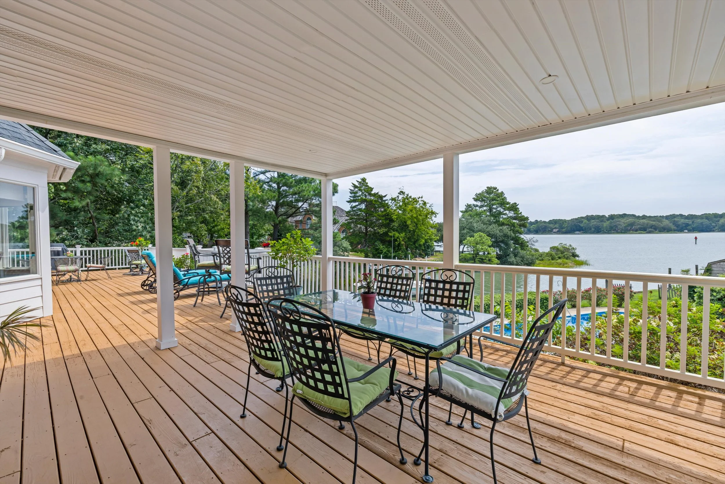 The image shows a spacious wooden deck with outdoor furniture, including a glass-top dining table with chairs and cushions. It is covered by a white ceiling with built-in lighting. The background features scenic views of a lake, trees, and a partly c