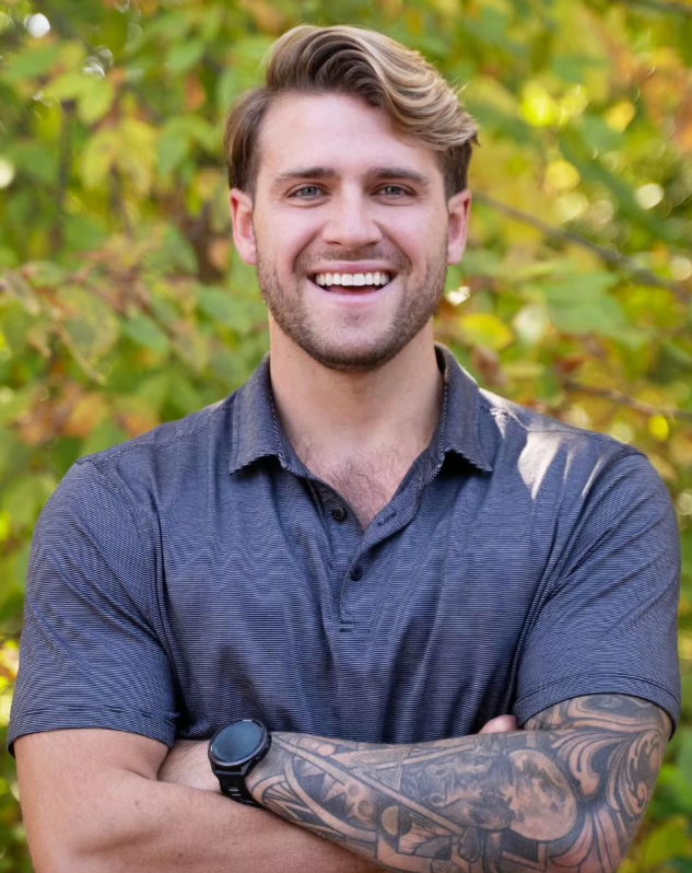 A smiling young man with light brown hair and a beard, wearing a dark blue polo shirt, standing outdoors with green leafy trees in the background.