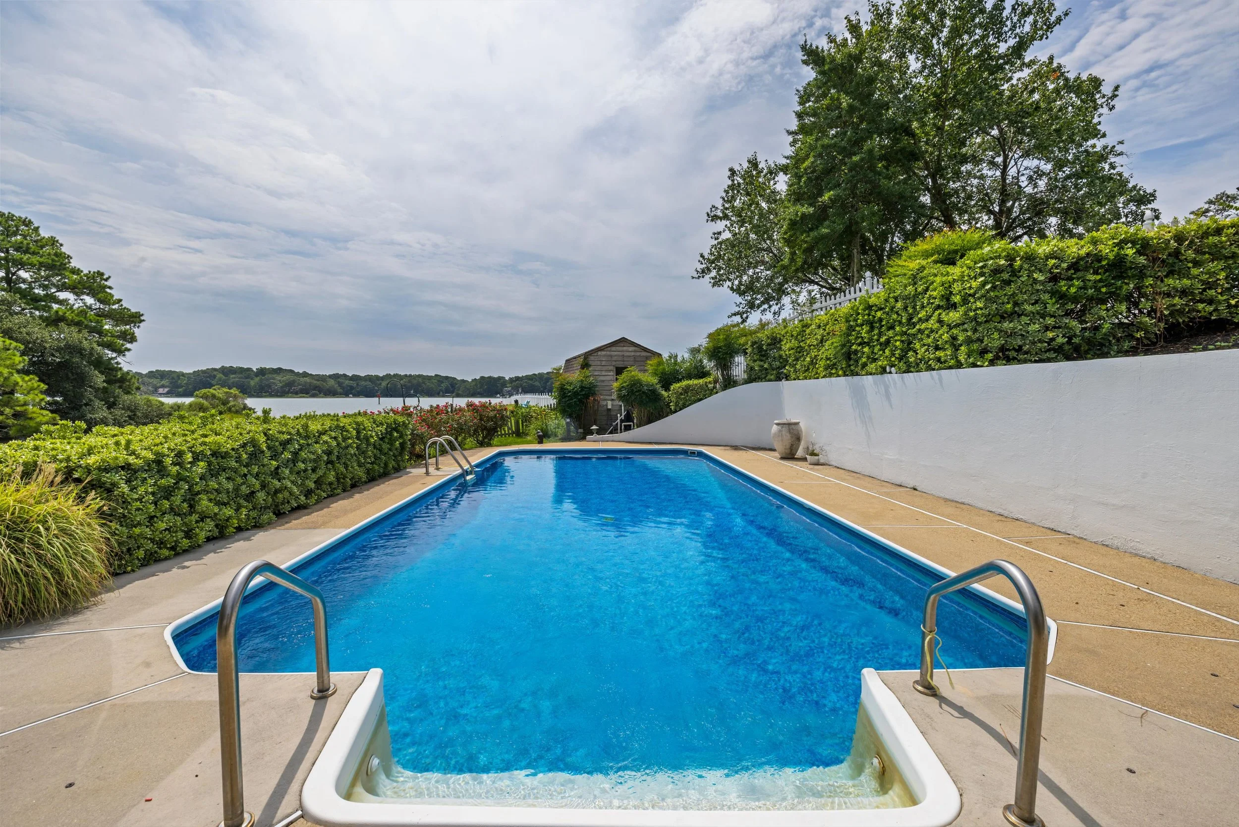 A backyard swimming pool with a scenic view of a lake and trees, surrounded by green bushes and a white fence, under a cloudy sky.