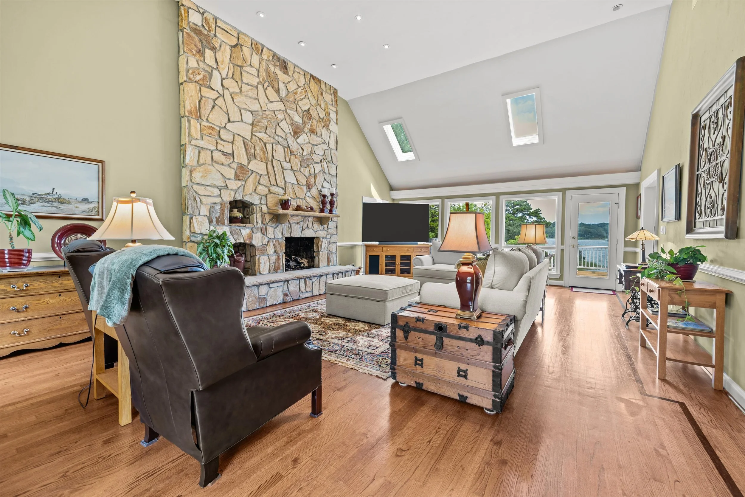 A cozy living room with a stone fireplace, beige sofas, wooden furniture, potted plants, and large windows with views of trees and water.