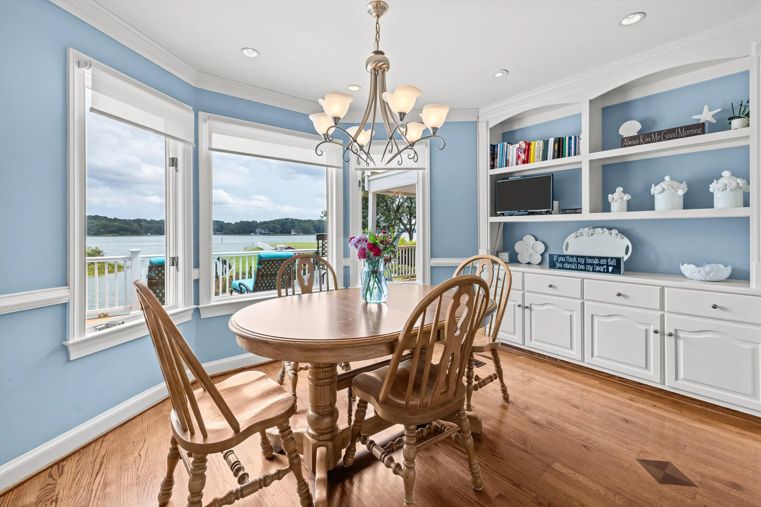 A dining room with a wooden table and matching chairs near large windows overlooking a waterfront landscape. The room has light blue walls, white crown molding, a chandelier with multiple light fixtures, and built-in white shelving with books and dec