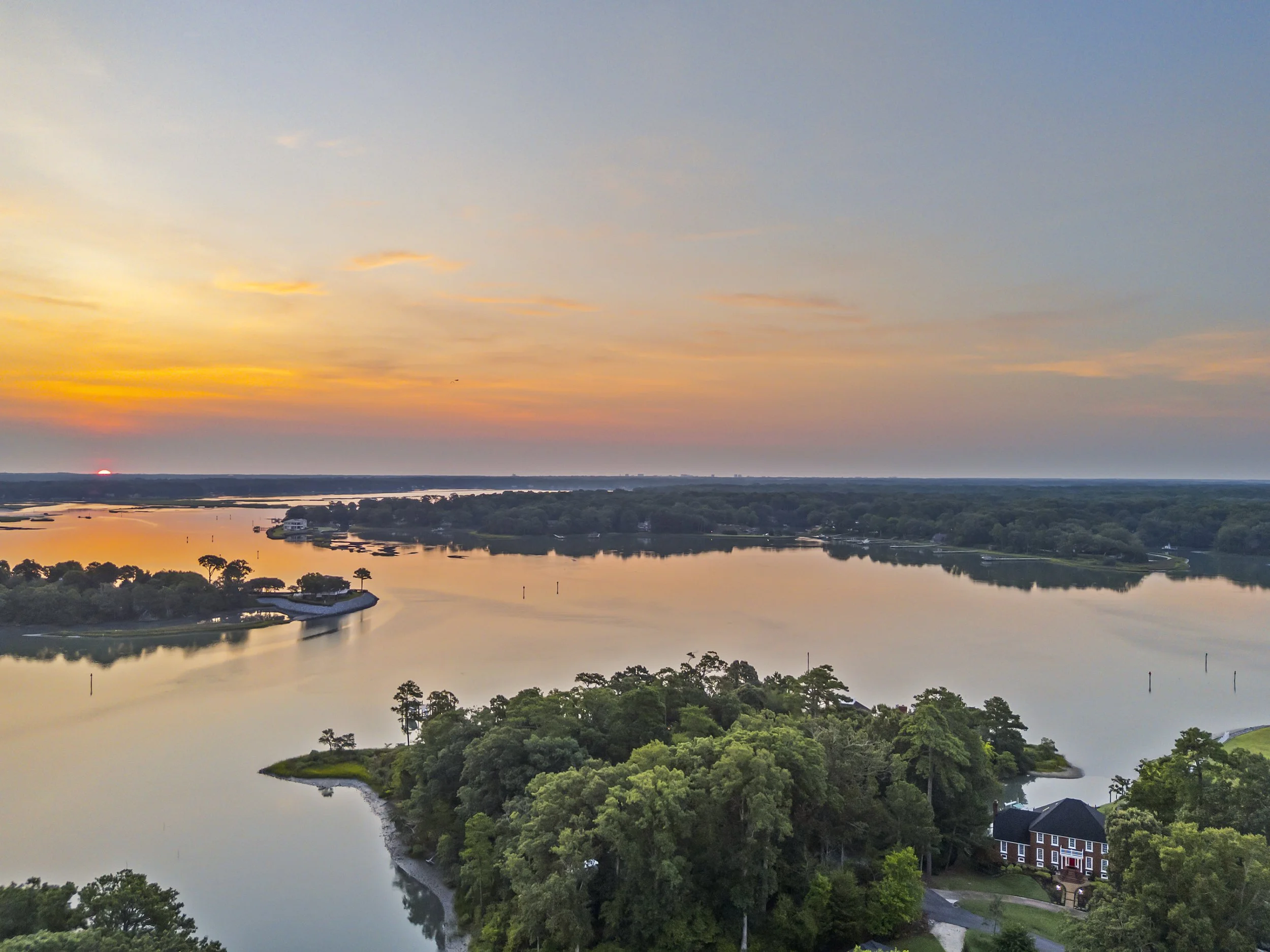 Sunset over a river with a tree-lined peninsula and residential houses.