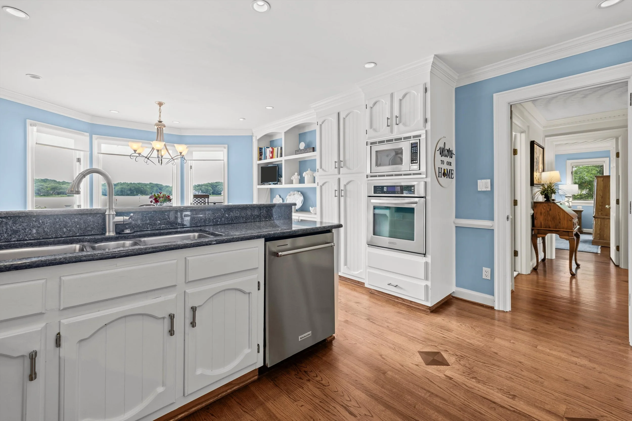 A spacious kitchen with white cabinets, black granite countertops, hardwood floors, and a view of a bright dining area with large windows and a chandelier.