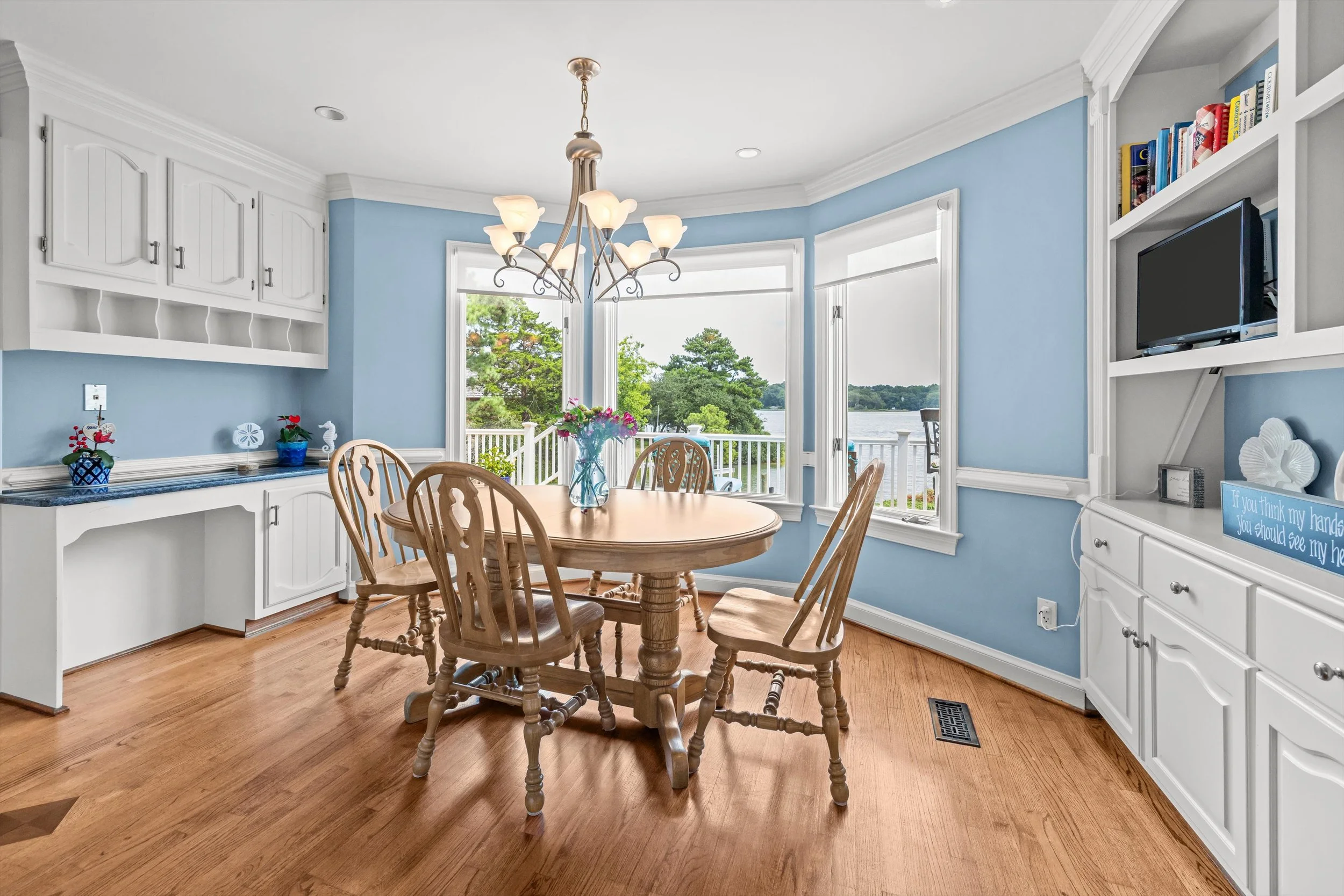 A dining area with a wooden table and six matching chairs, large windows with a view of trees and water, white built-in cabinets, a hanging chandelier, and a bookshelf with books and decorations.