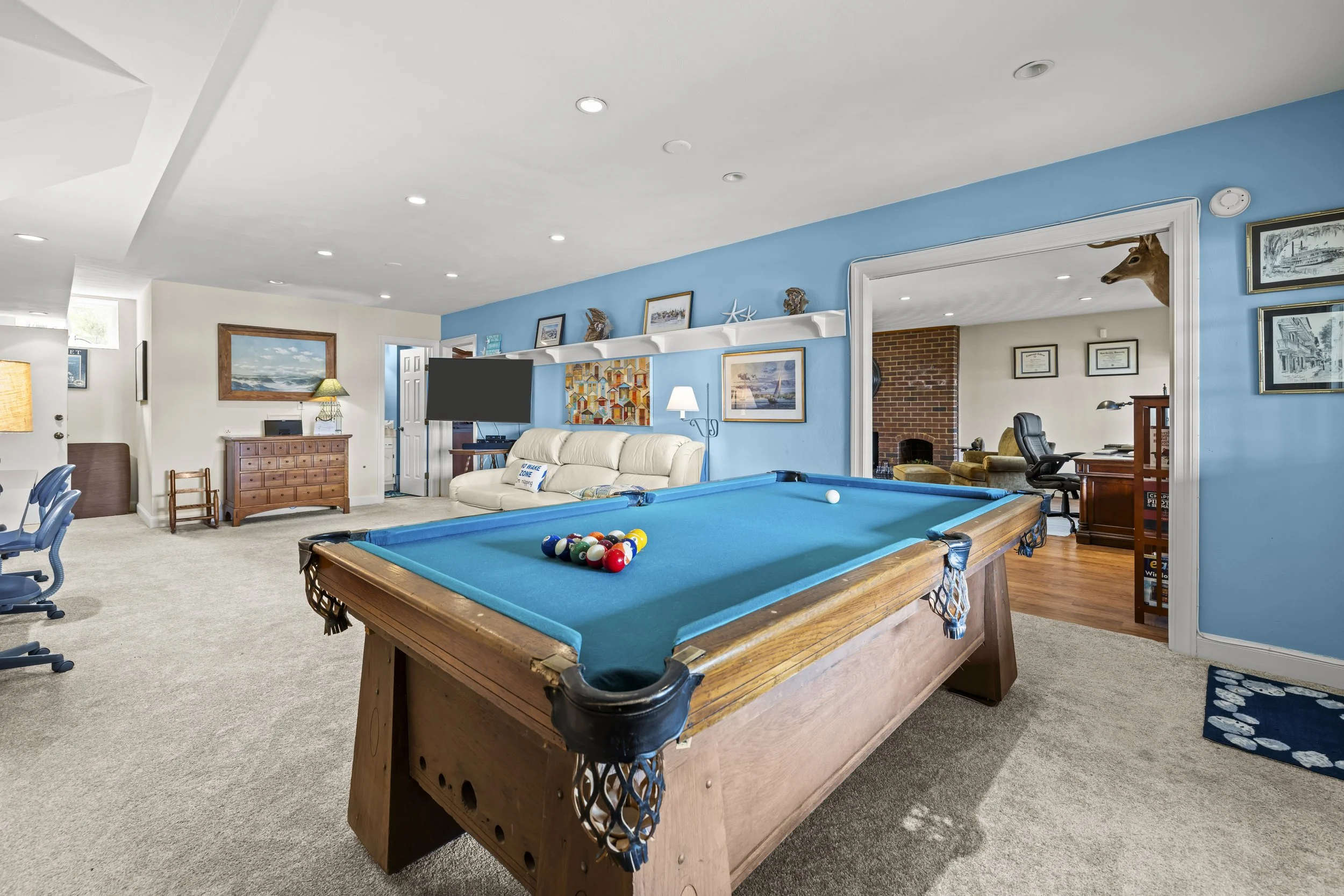 A basement game room with a pool table in the foreground and a white couch behind it. The room is decorated with coastal artwork and nautical objects on a blue accent wall. There is a flat-screen TV and a wooden dresser on the left side. An open door