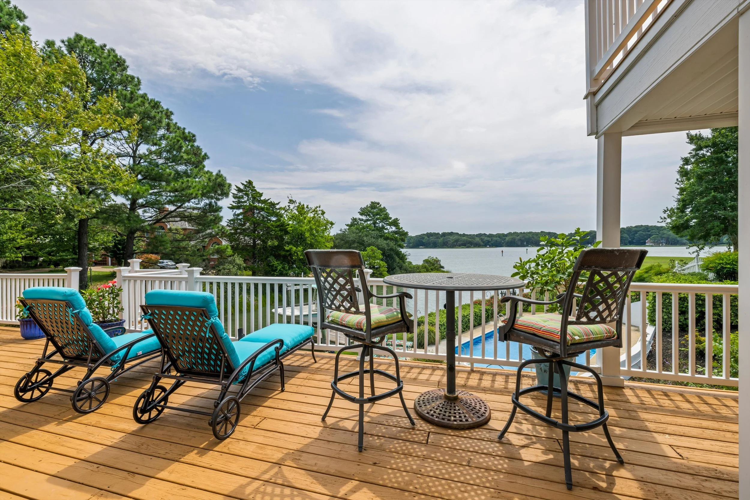 A wooden deck with outdoor furniture, including a round table, chairs with cushions, and a lounge chair. The deck overlooks a lake with trees and a partly cloudy sky in the background.
