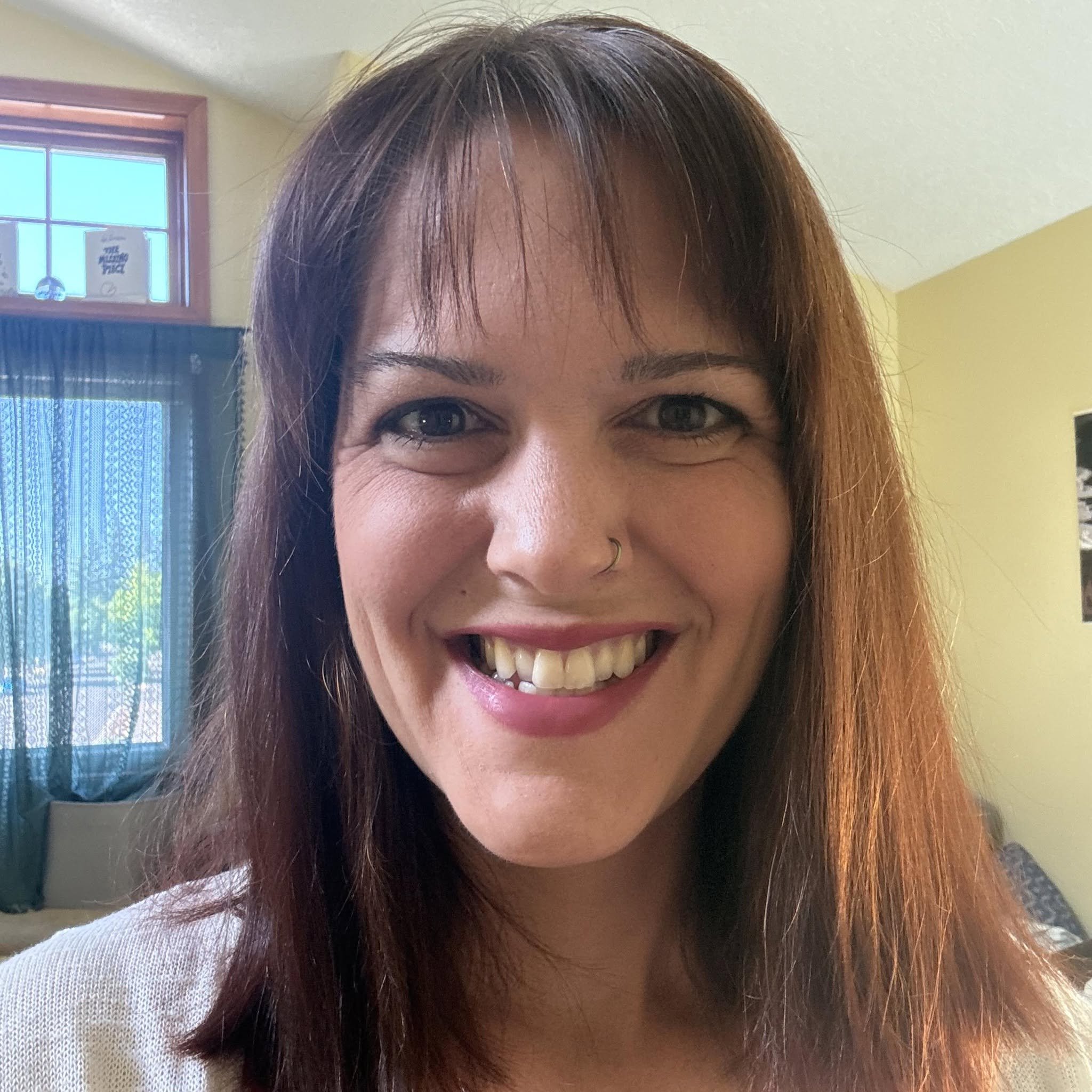 Close-up of a smiling woman's face with brown hair and dark eyes, indoors near a window with a plant and decorative items in the background.