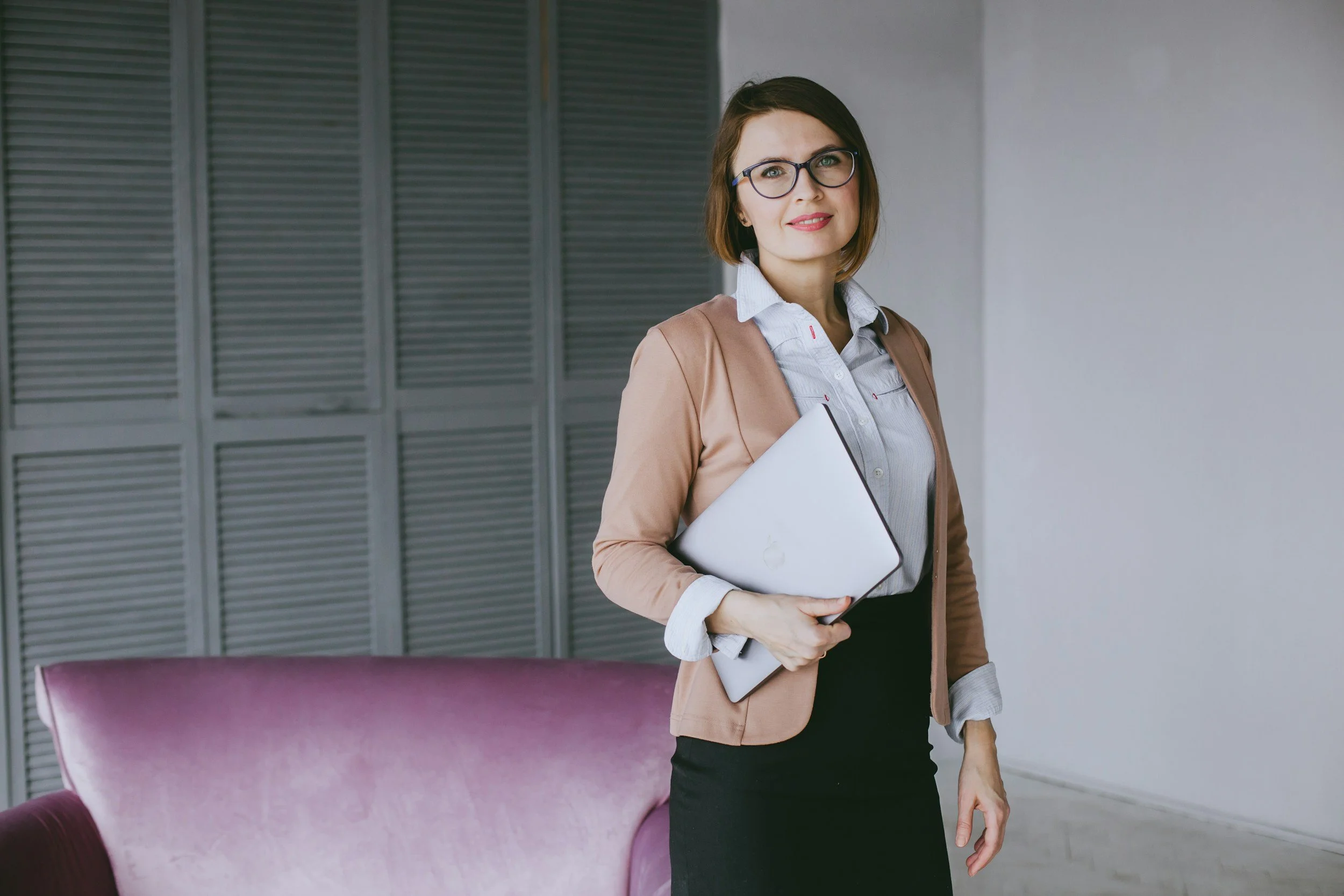 A woman with glasses, wearing a light gray shirt and a brown blazer, holding a closed laptop and standing in front of a gray louvered closet door and a purple cushioned chair.