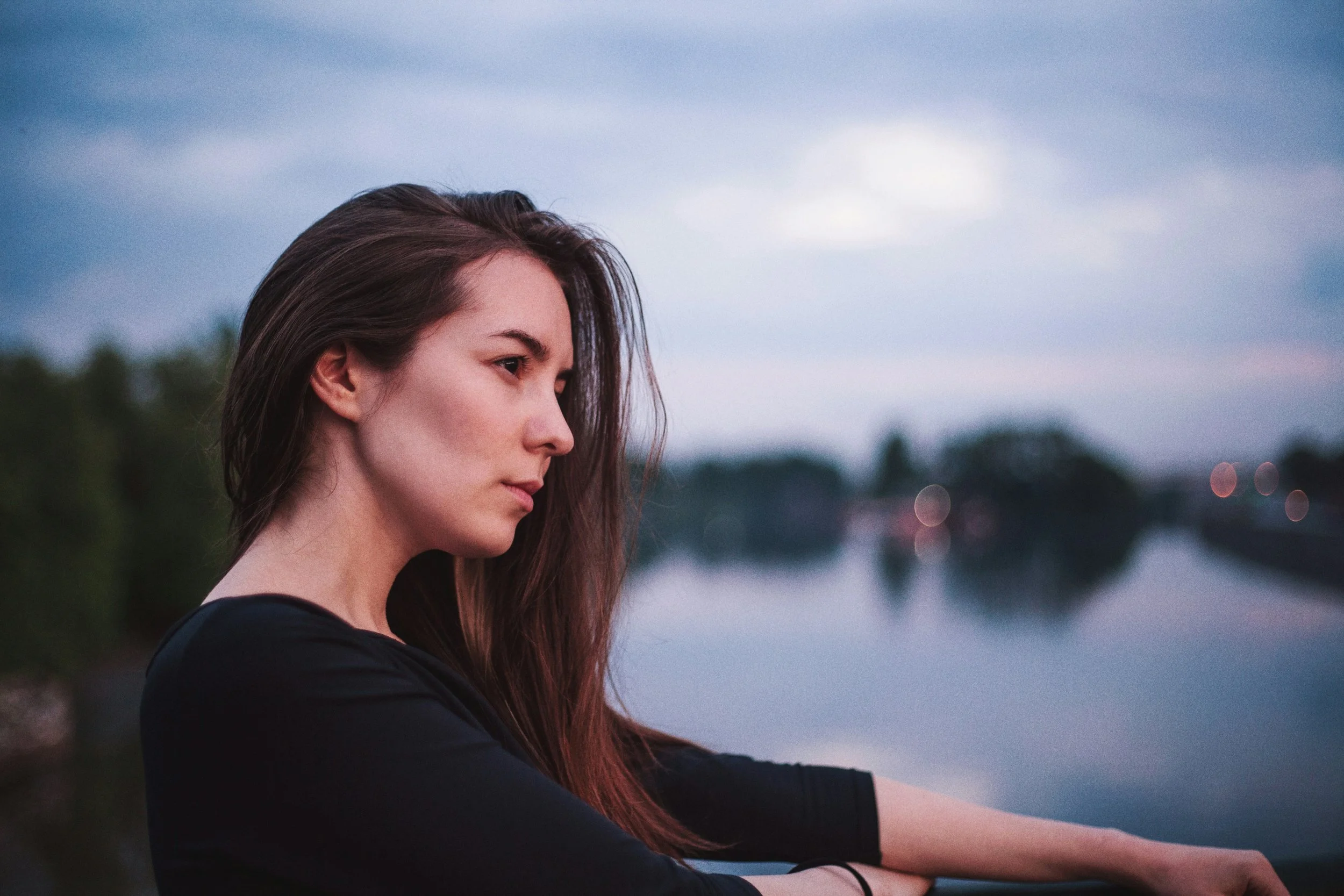 Side profile of a young woman with long brown hair looking at dusk or dawn near a body of water with blurry trees and lights in the background.
