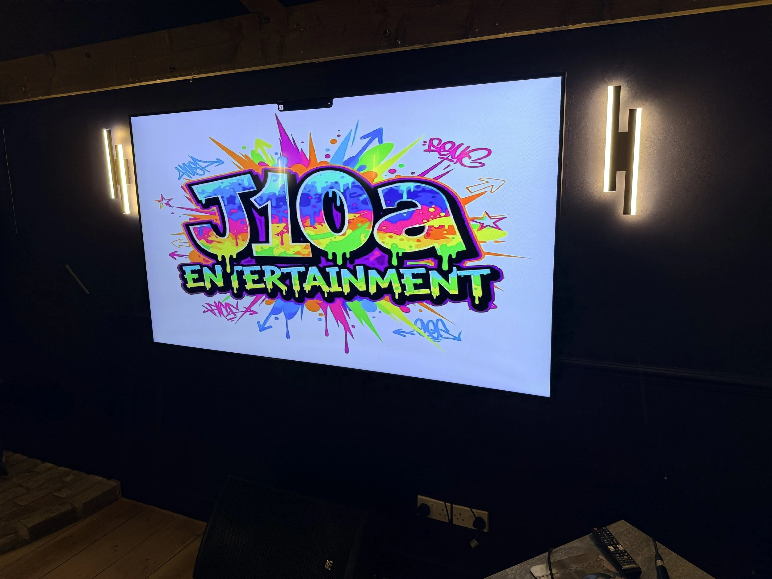 A large digital screen displaying colorful graffiti-style text that reads "105 ENTERTAINMENT," with vibrant splashes and arrows around it, mounted on a dark wall with wall-mounted lights on each side.