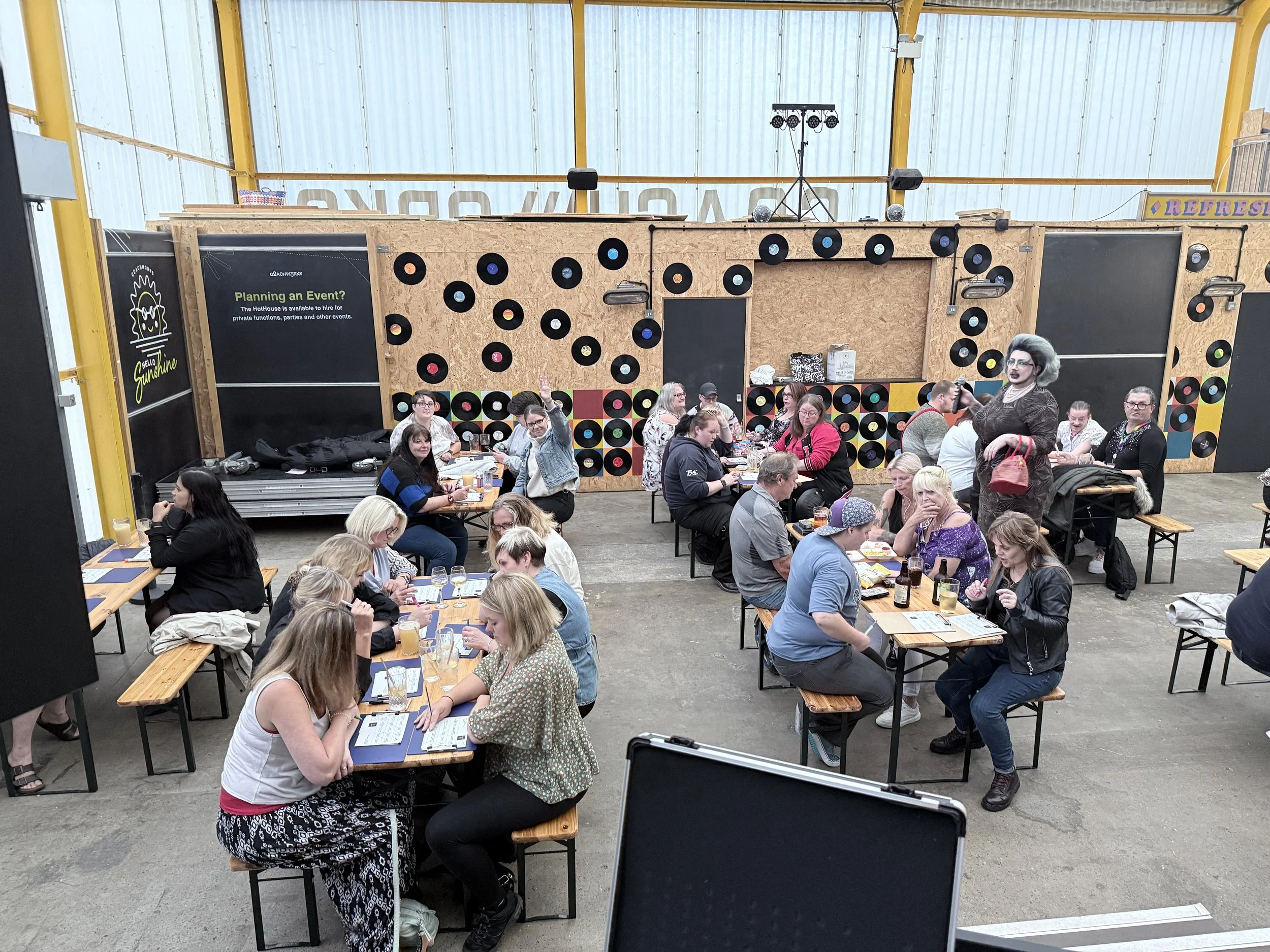 People sitting at picnic tables inside a large industrial-style venue with wooden walls decorated with vinyl records, and a menu board reading "Planning an Event?".