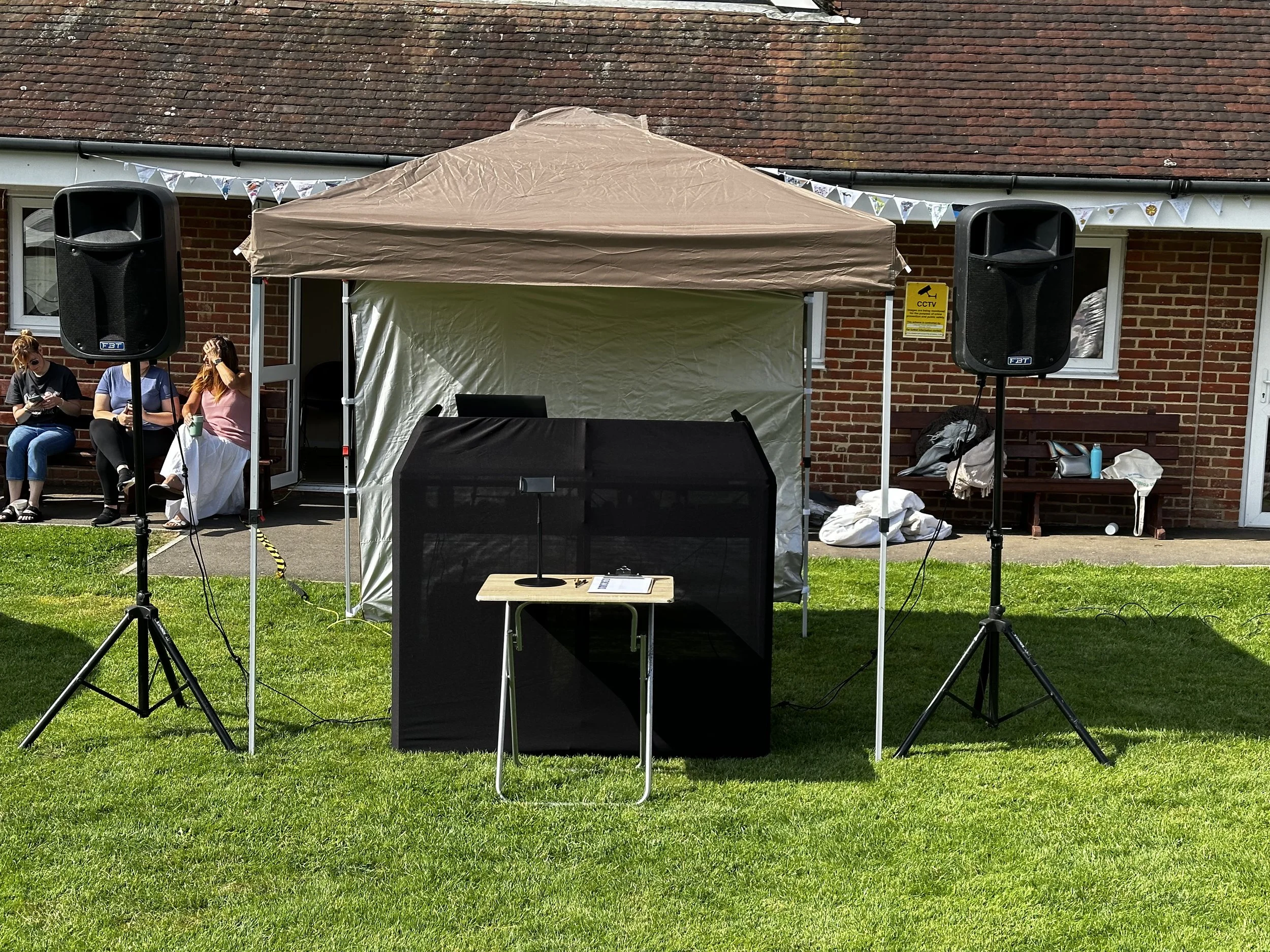 An outdoor event setup in front of a brick house with a pitched roof. There is a beige canopy tent, two large black speakers on stands, and people sitting on a bench. Decorations include bunting flags, and there are various items like bags, water bot