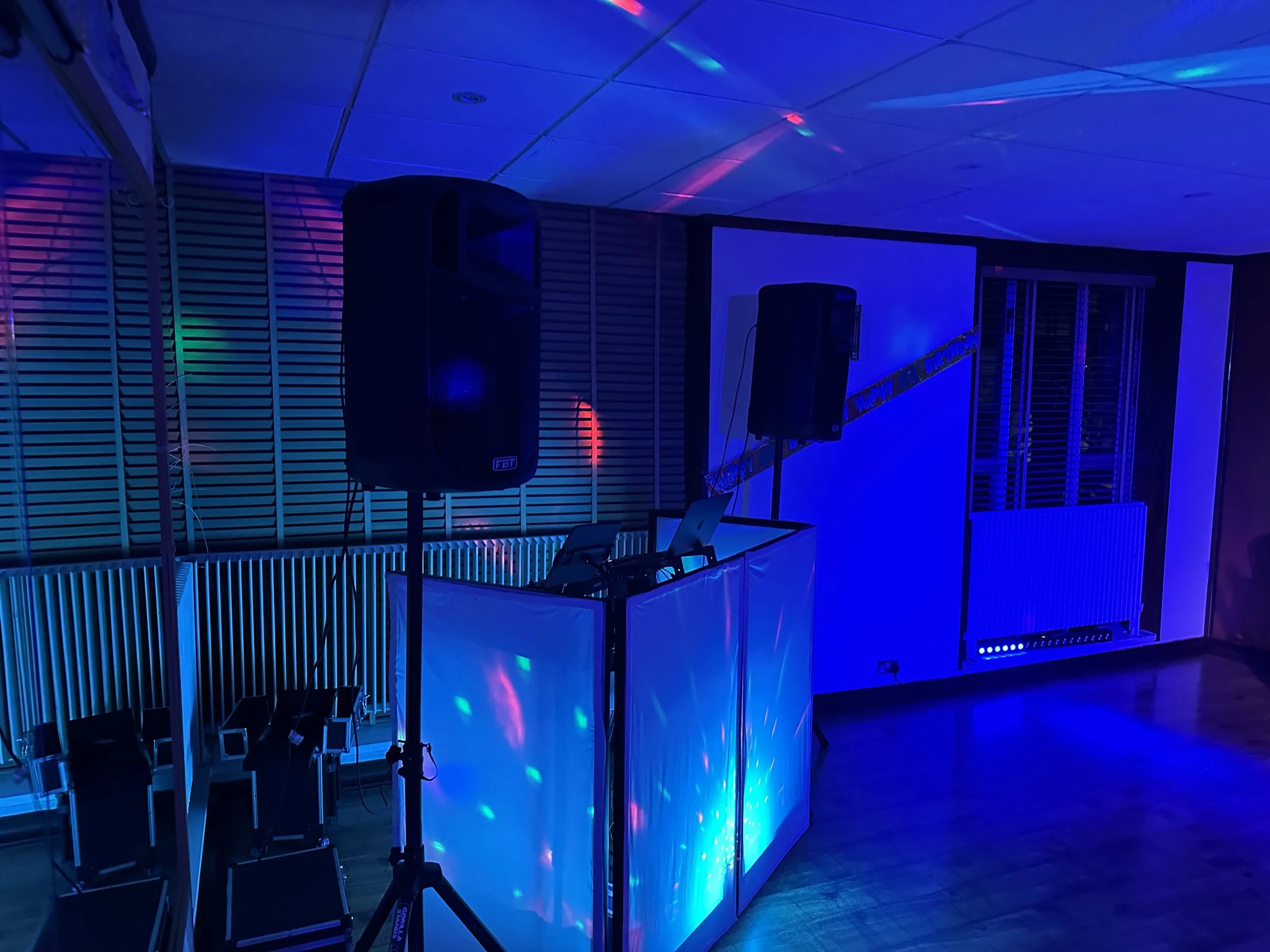 A dark room decorated with colorful laser lights, featuring a DJ booth with speakers and a laptop, and rows of chairs along the wall, suggesting a party or celebration setting.